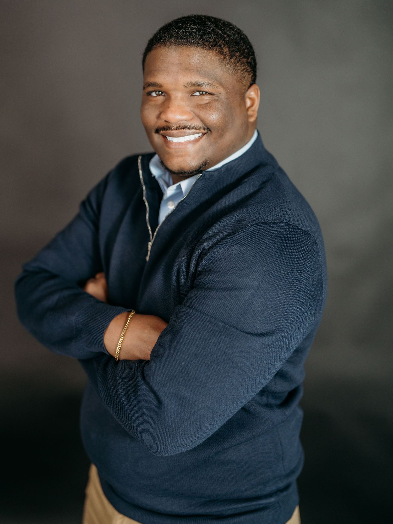 Professional business portrait of a person smiling warmly while wearing a navy blue sweater against a dark backdrop.