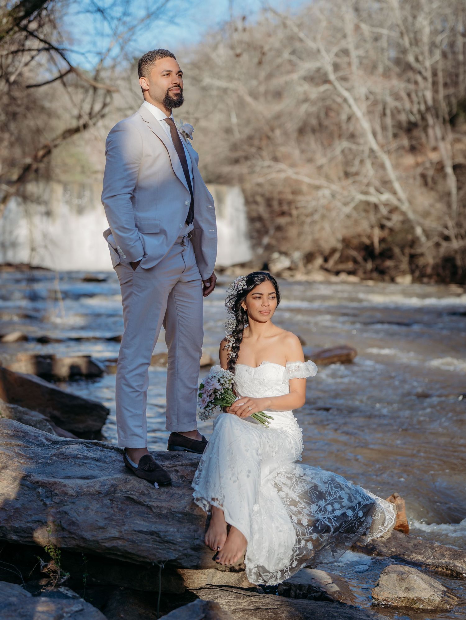 A couple shares romantic moments by the rocky riverbank with bare winter trees in the background.