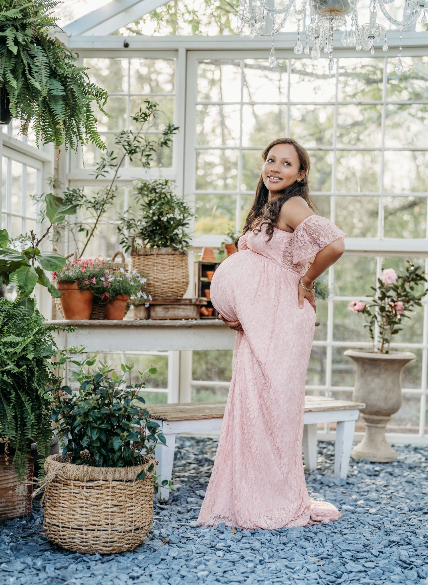 Elegant maternity photo of woman in blush lace gown standing among potted plants in glass conservatory setting.