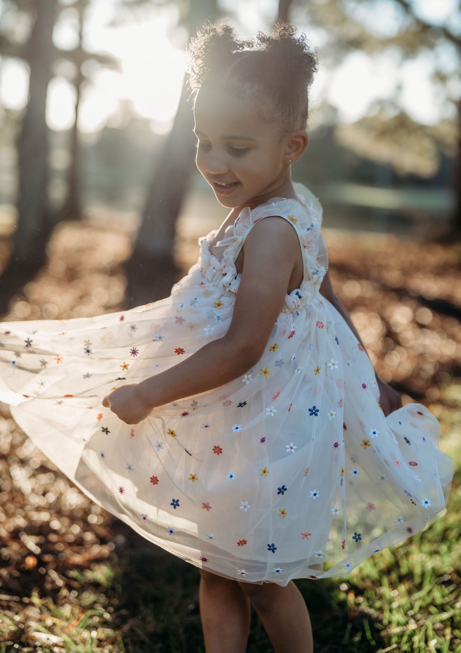 A child in a white floral dress twirls in dappled sunlight among autumn leaves on the ground.