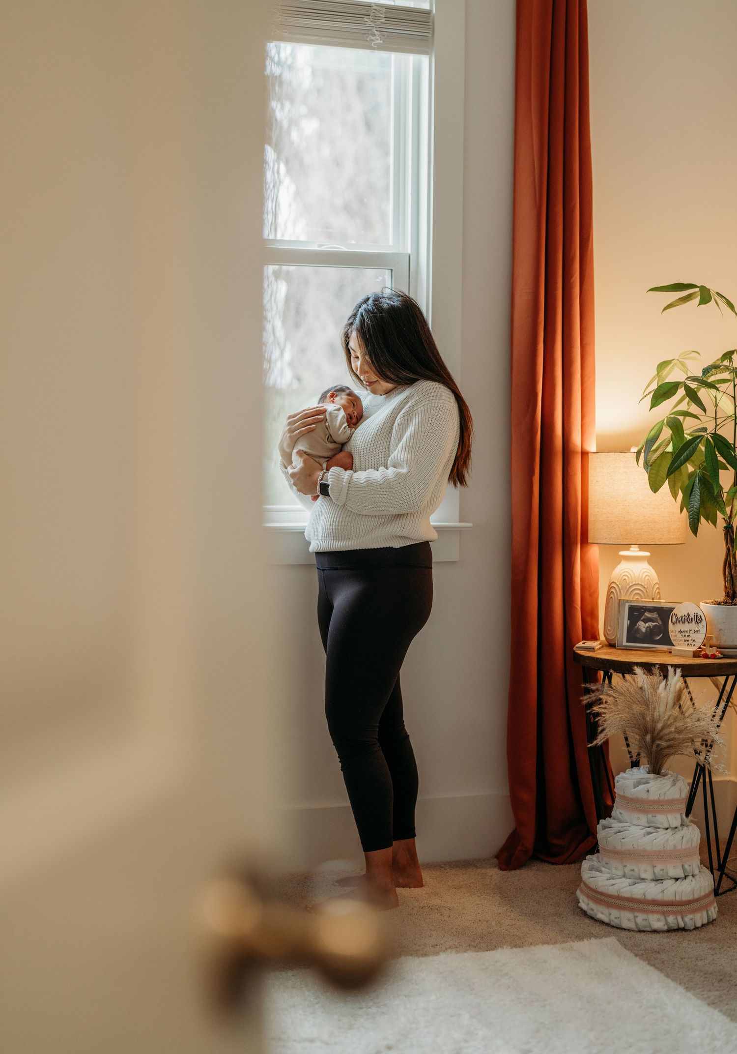 Interior scene with white diaper cake on table near window with orange curtains and house plant in soft natural lighting.