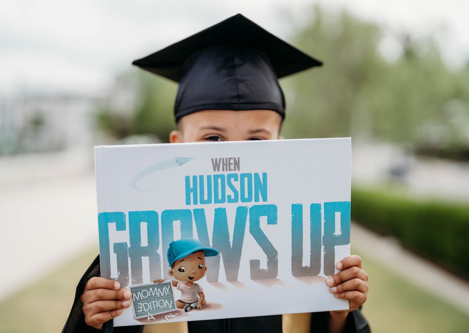 Child in graduation cap holds personalized book titled When Hudson Grows Up, celebrating academic milestone.