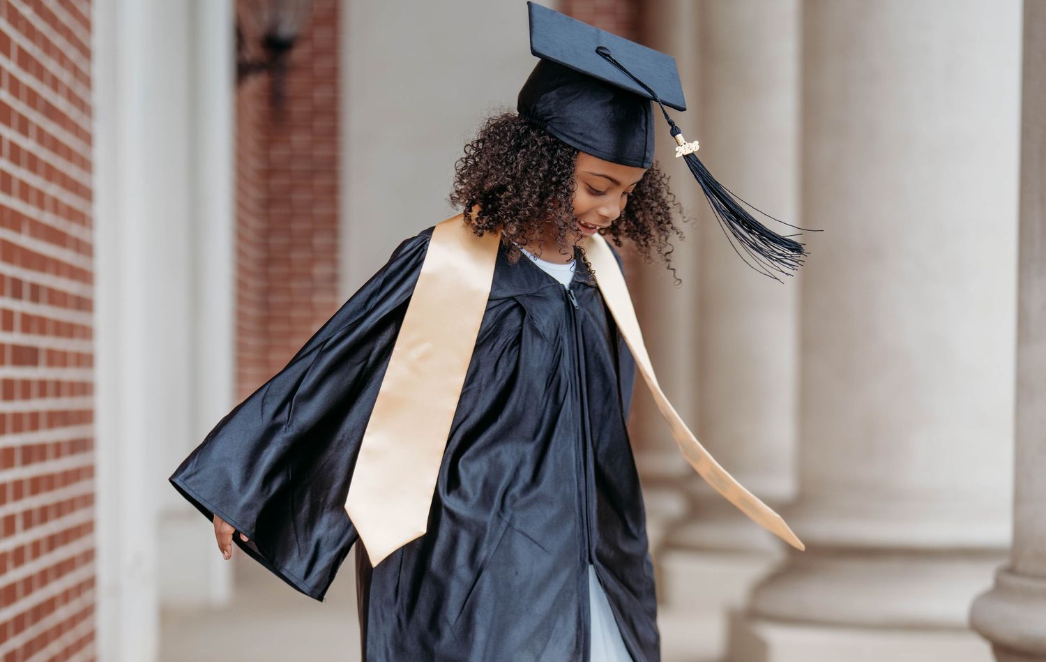Young woman in black graduation gown and cap with gold sash celebrates her achievement outdoors near brick columns.