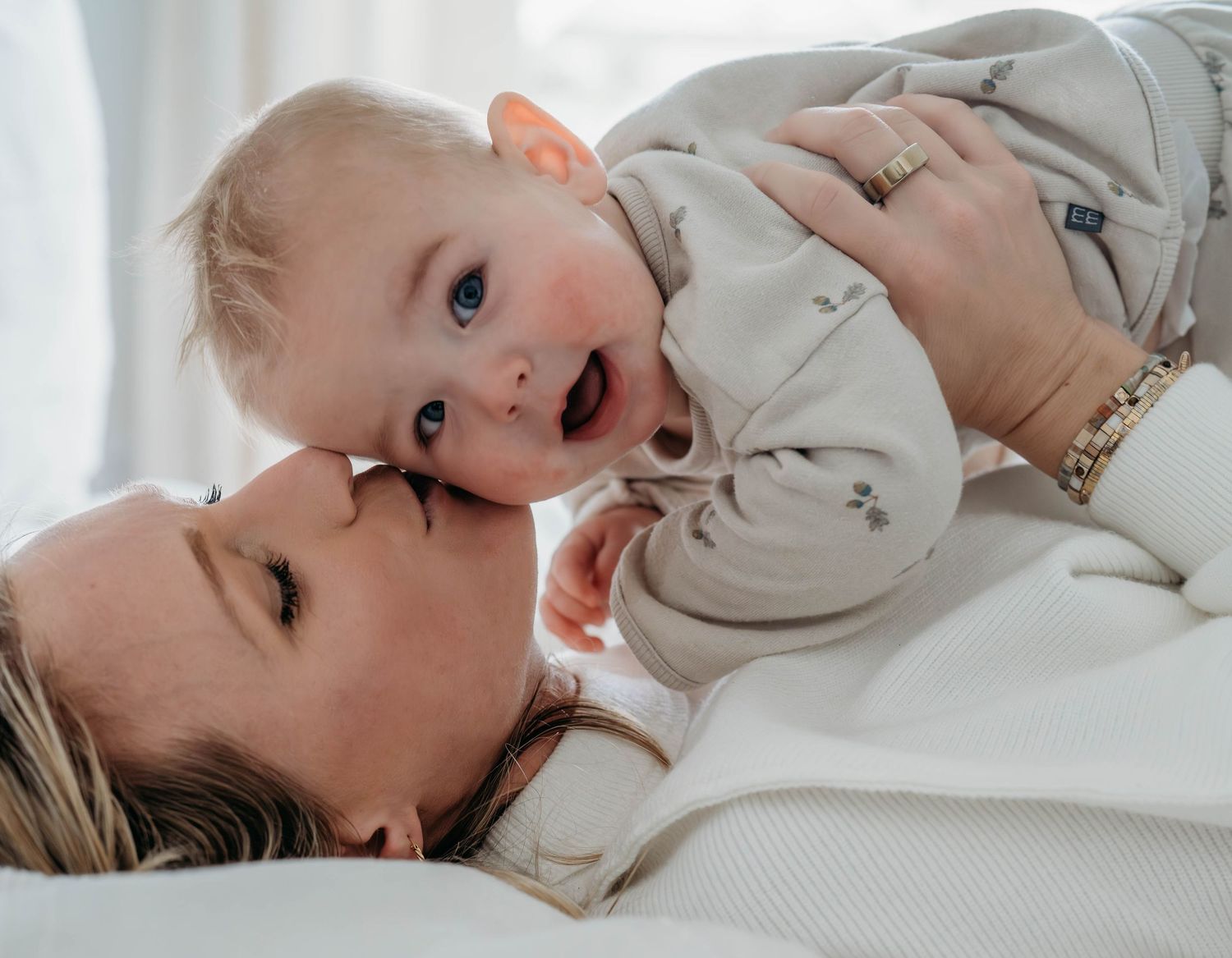 A happy baby in a light-colored onesie being held by mother, while smiling and playing on a white bed setting