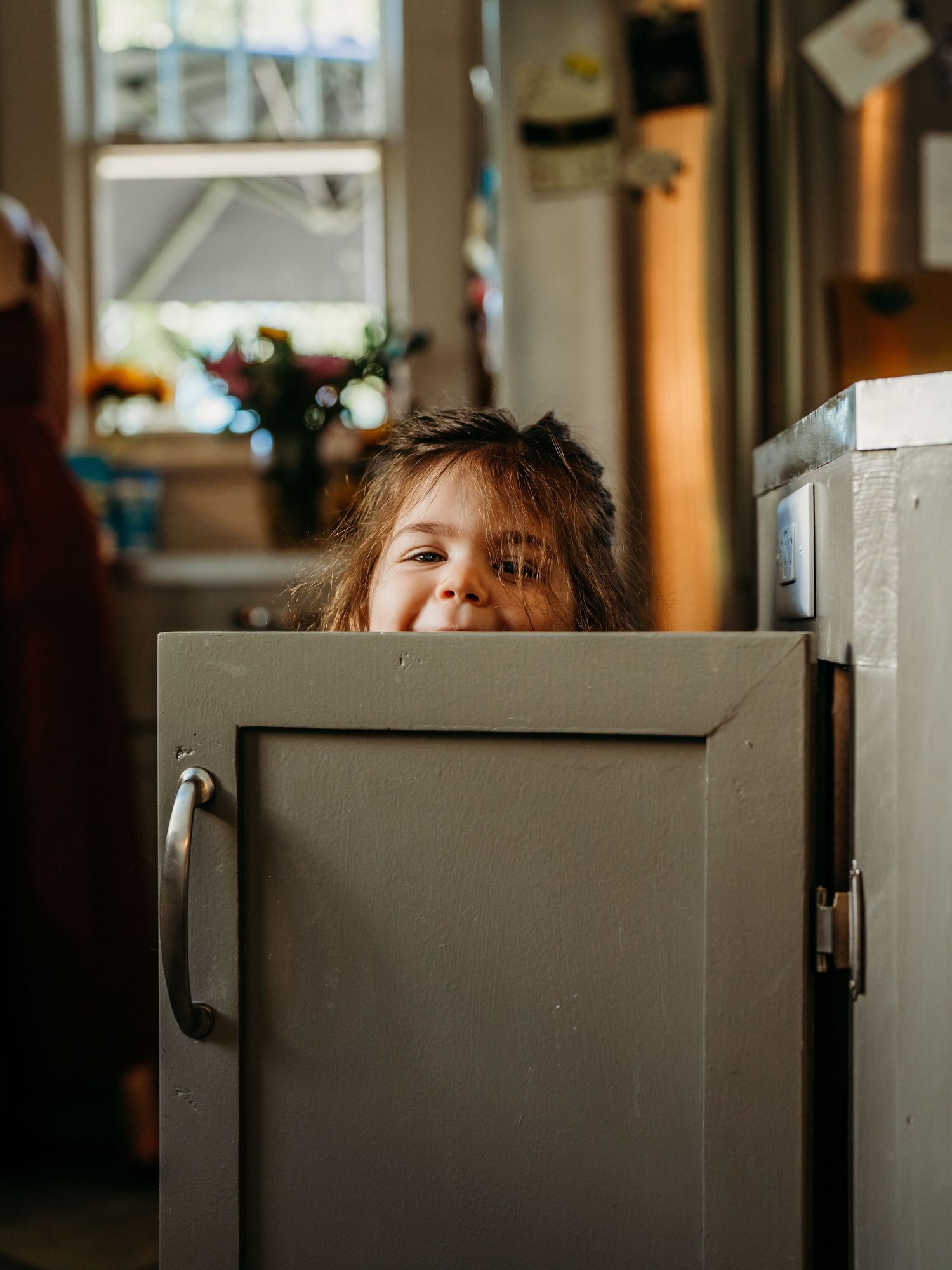 A playful moment captured as a child peeks out from behind a gray cabinet door in warm indoor lighting.