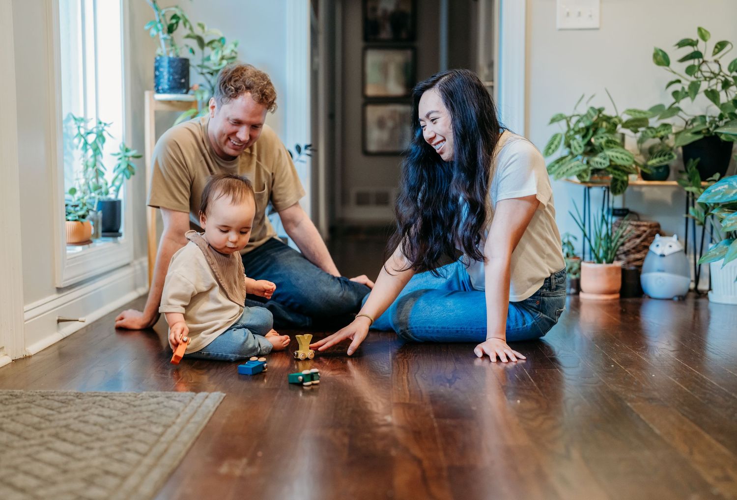 Family sits together on hardwood floor of home playing with toys surrounded by houseplants near window.