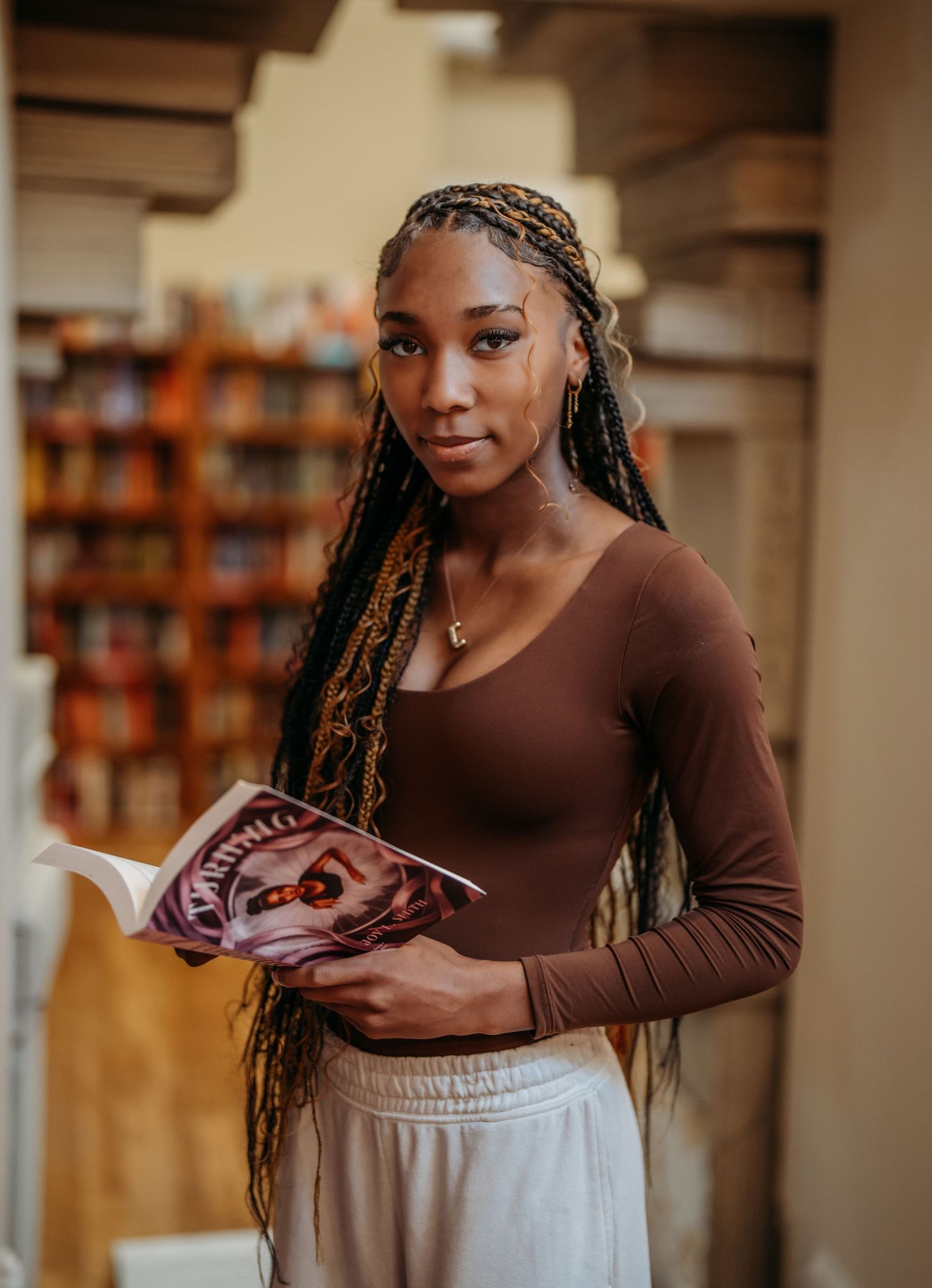 Student reading a magazine in a library while wearing a brown long sleeve shirt and beige pants with braided hair.