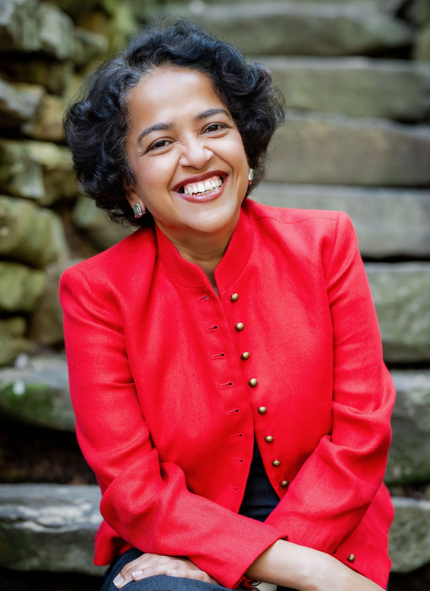 A person in a bright red blazer jacket poses against a stone wall background.