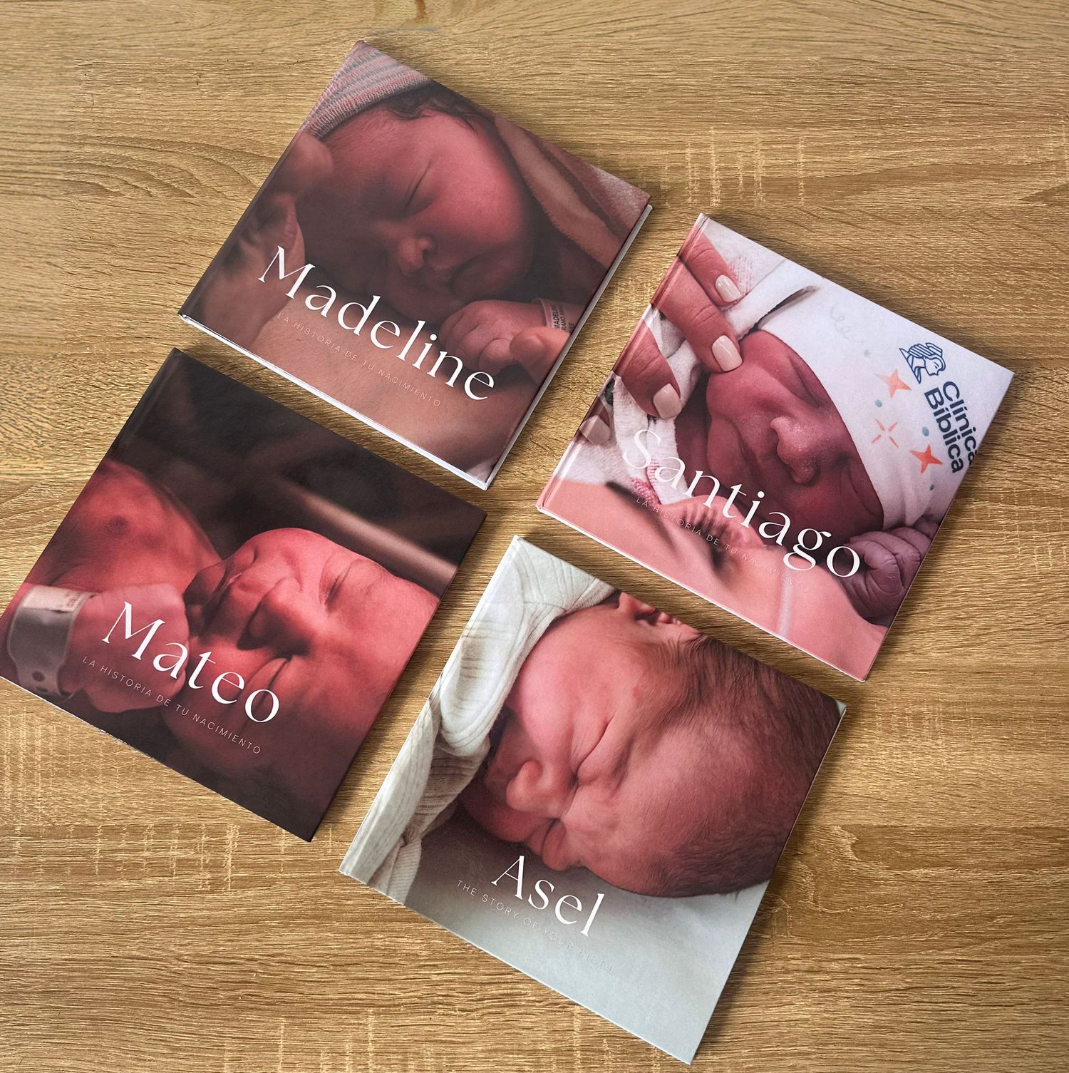 Four personalized newborn baby photo books named Madeline, Santiago, Mateo, and Asel displayed on a wooden surface.