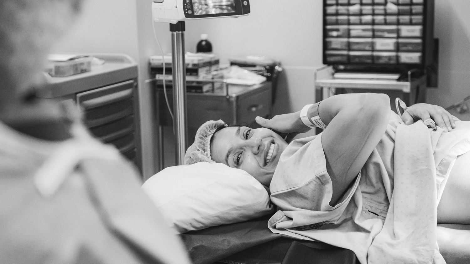 Black and white photo of a smiling woman lying in a hospital bed wearing a hair net during labor.