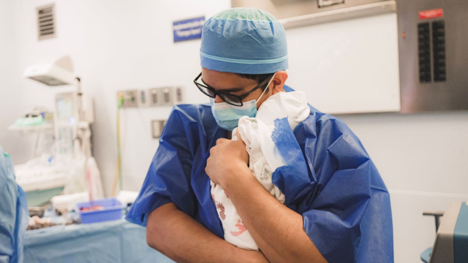A surgeon in blue scrubs and mask holds a newborn baby close to their chest in an operating room.