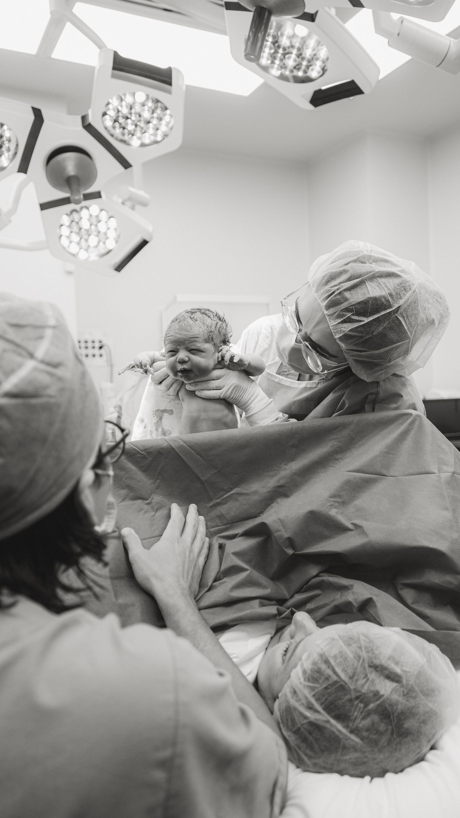 Black and white photo of a newborn baby being delivered by medical staff in an operating room under surgical lights.