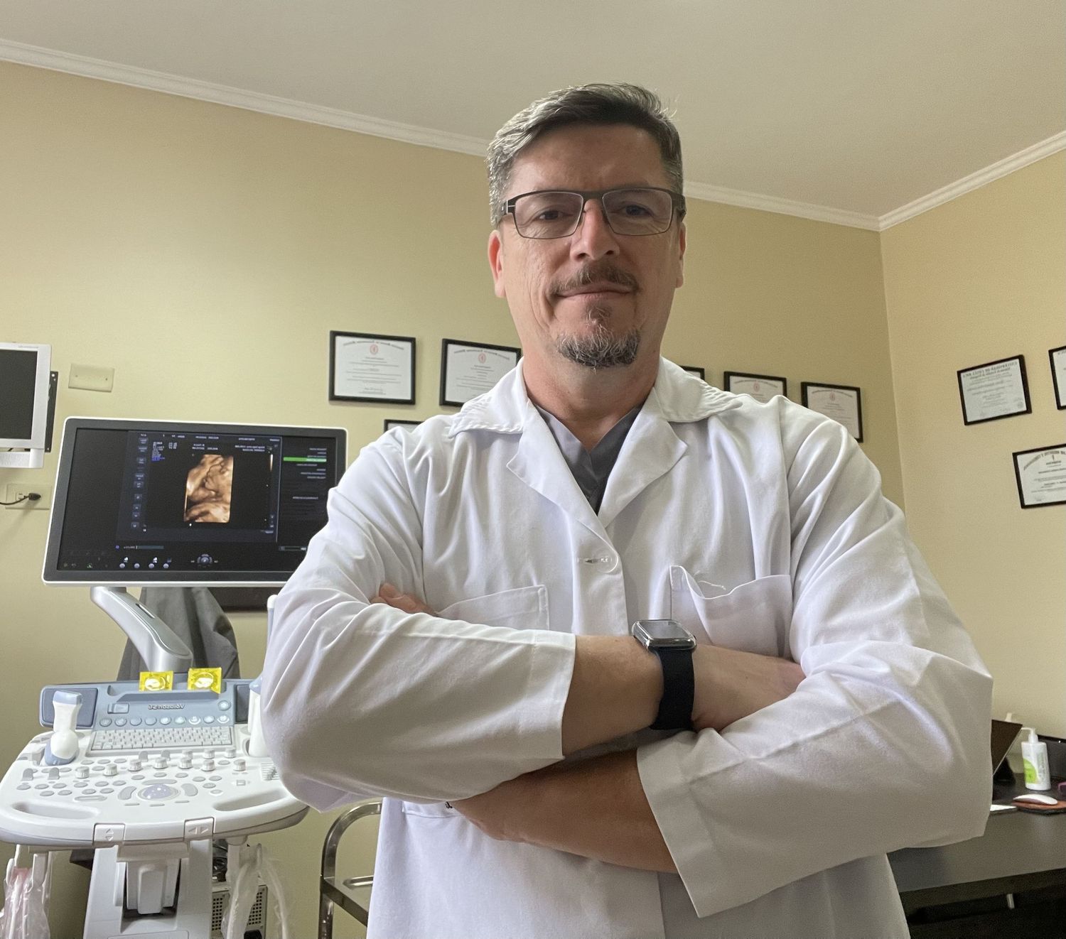 Doctor in white coat stands with crossed arms next to ultrasound equipment in medical office.