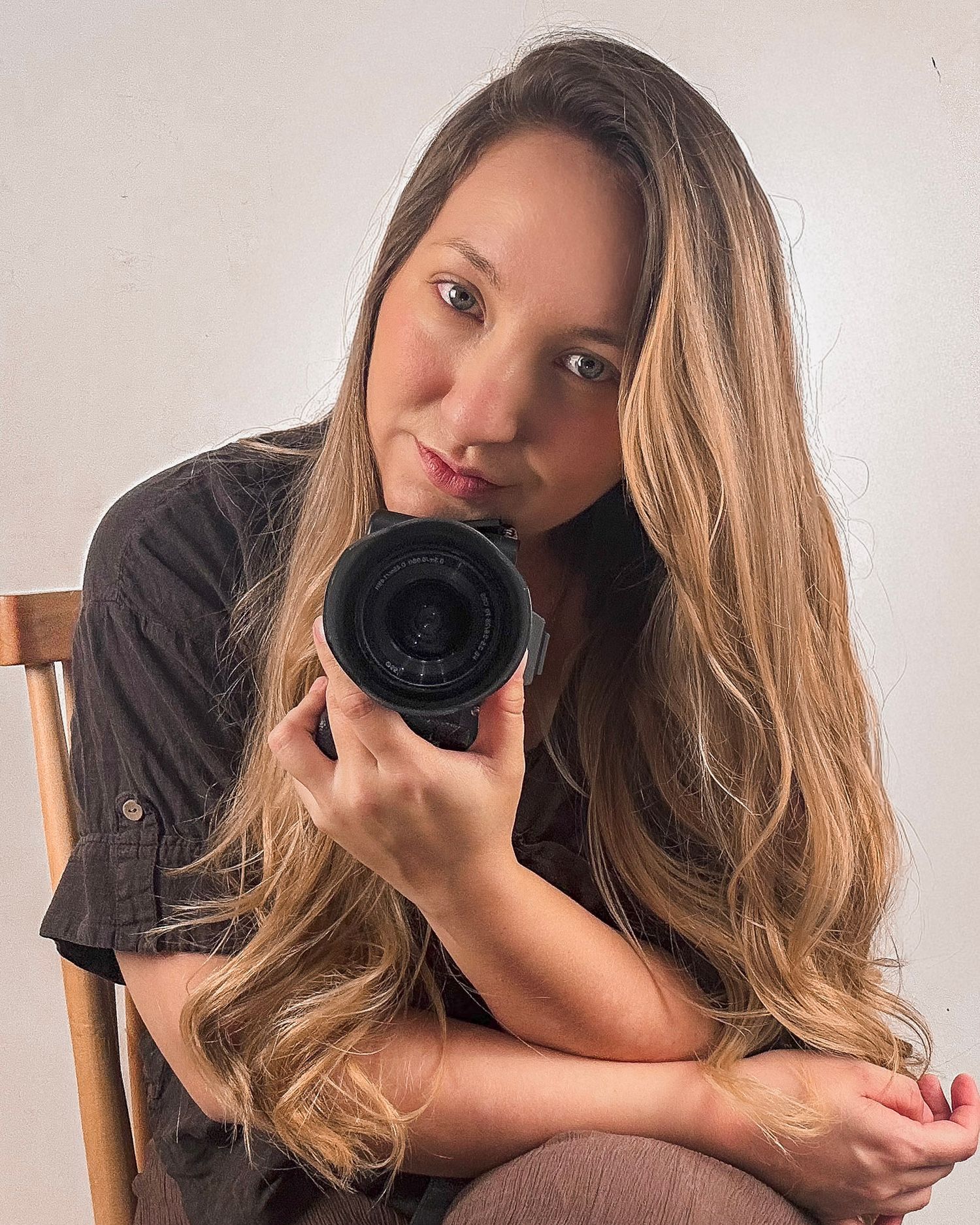 Young woman with long wavy hair holds a DSLR camera while sitting in a wooden chair against a white background.