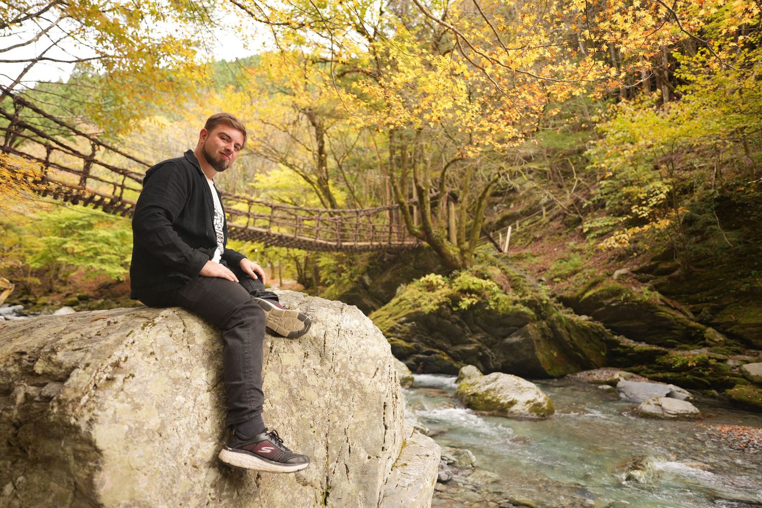 Scenic stream flowing through autumn forest with colorful fall foliage and person sitting on large rock.