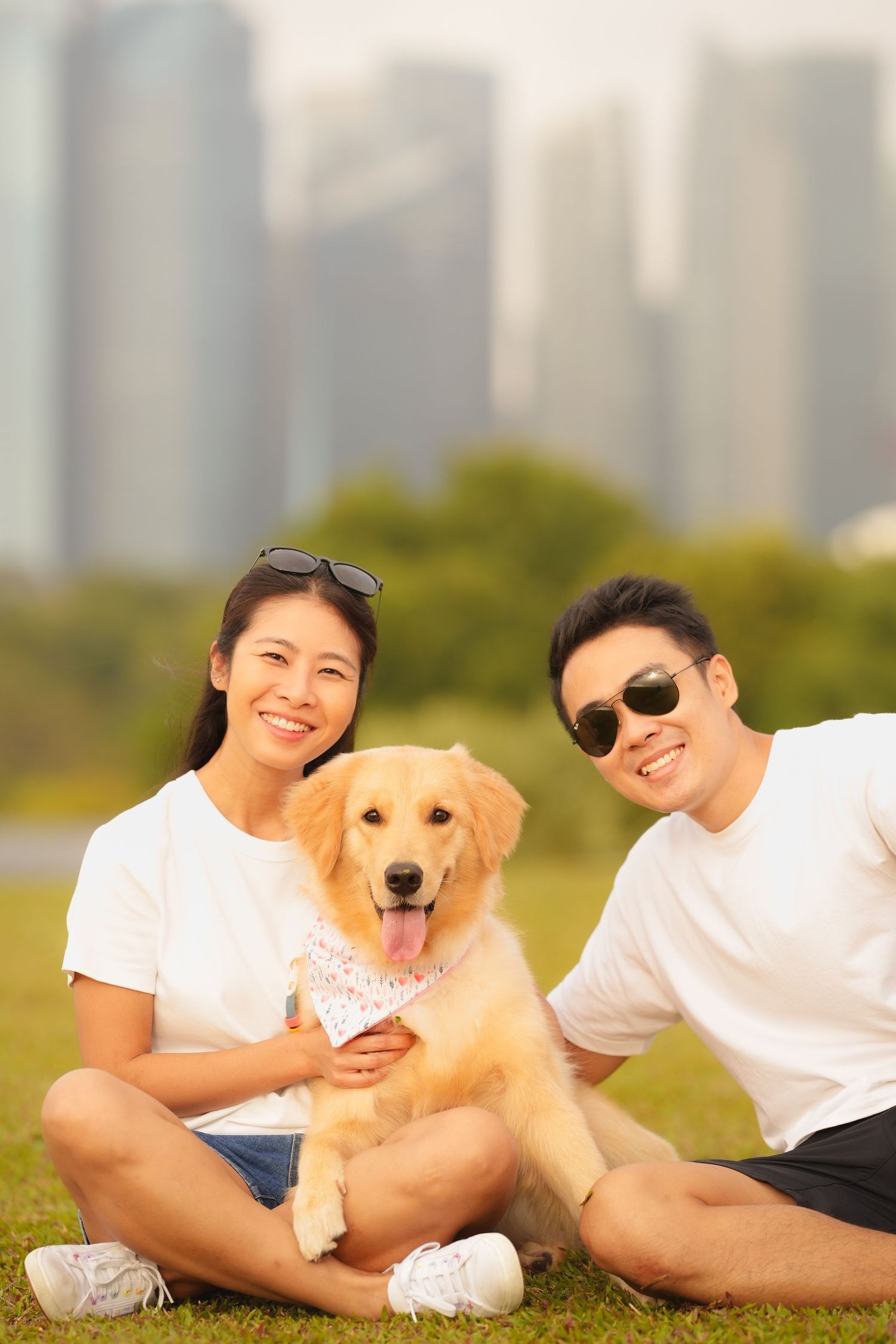 Two people sitting on grass with a smiling Golden Retriever puppy in a park with city skyline in background.