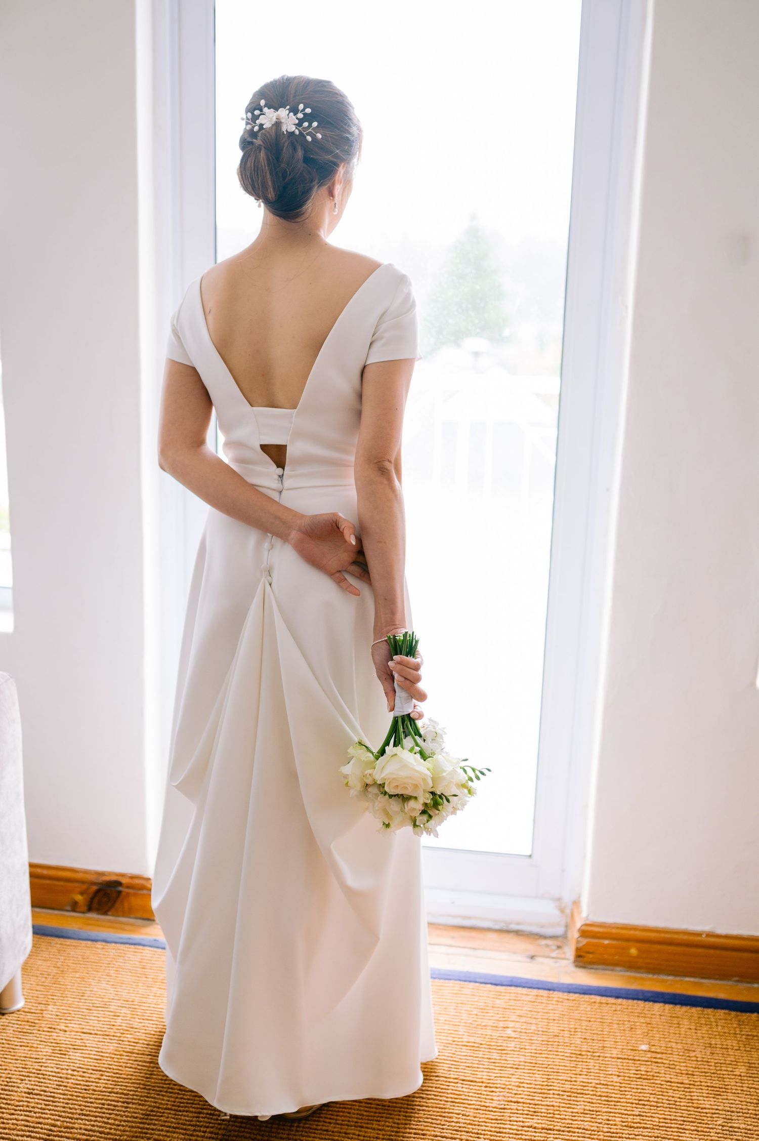 A bride in a white wedding dress holds a bouquet while standing by bright windows in a sunlit room.
