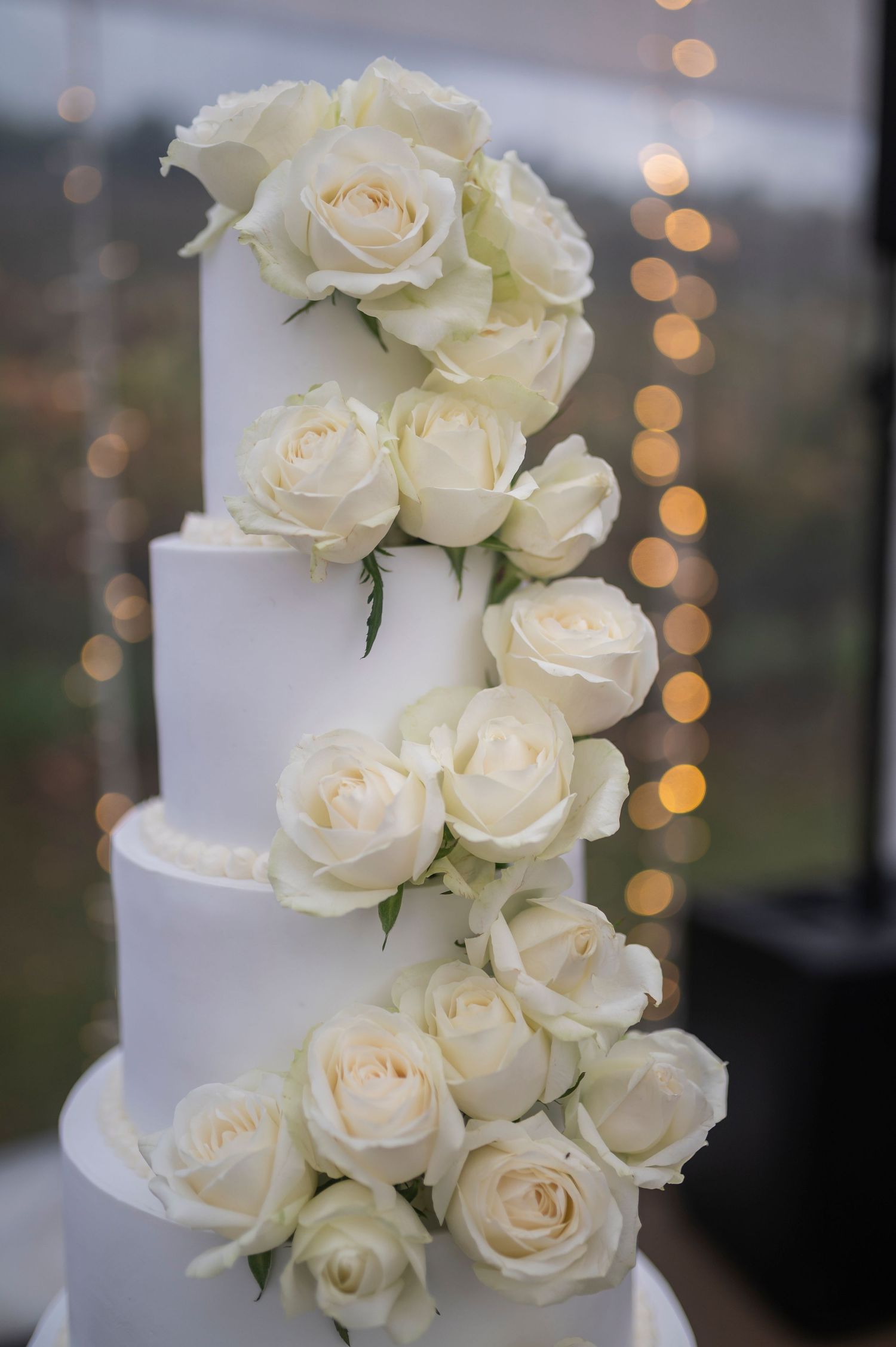 Elegant white wedding cake decorated with cascading cream roses against a bokeh light background.