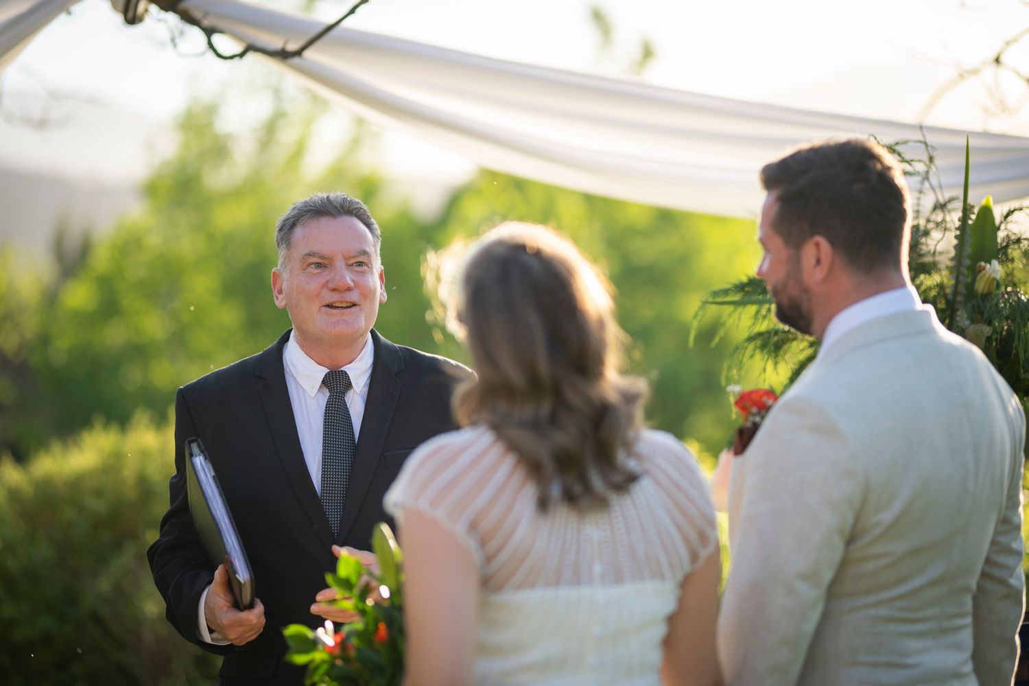 A group of guests celebrate at an outdoor wedding reception with bright sunlight filtering through trees.