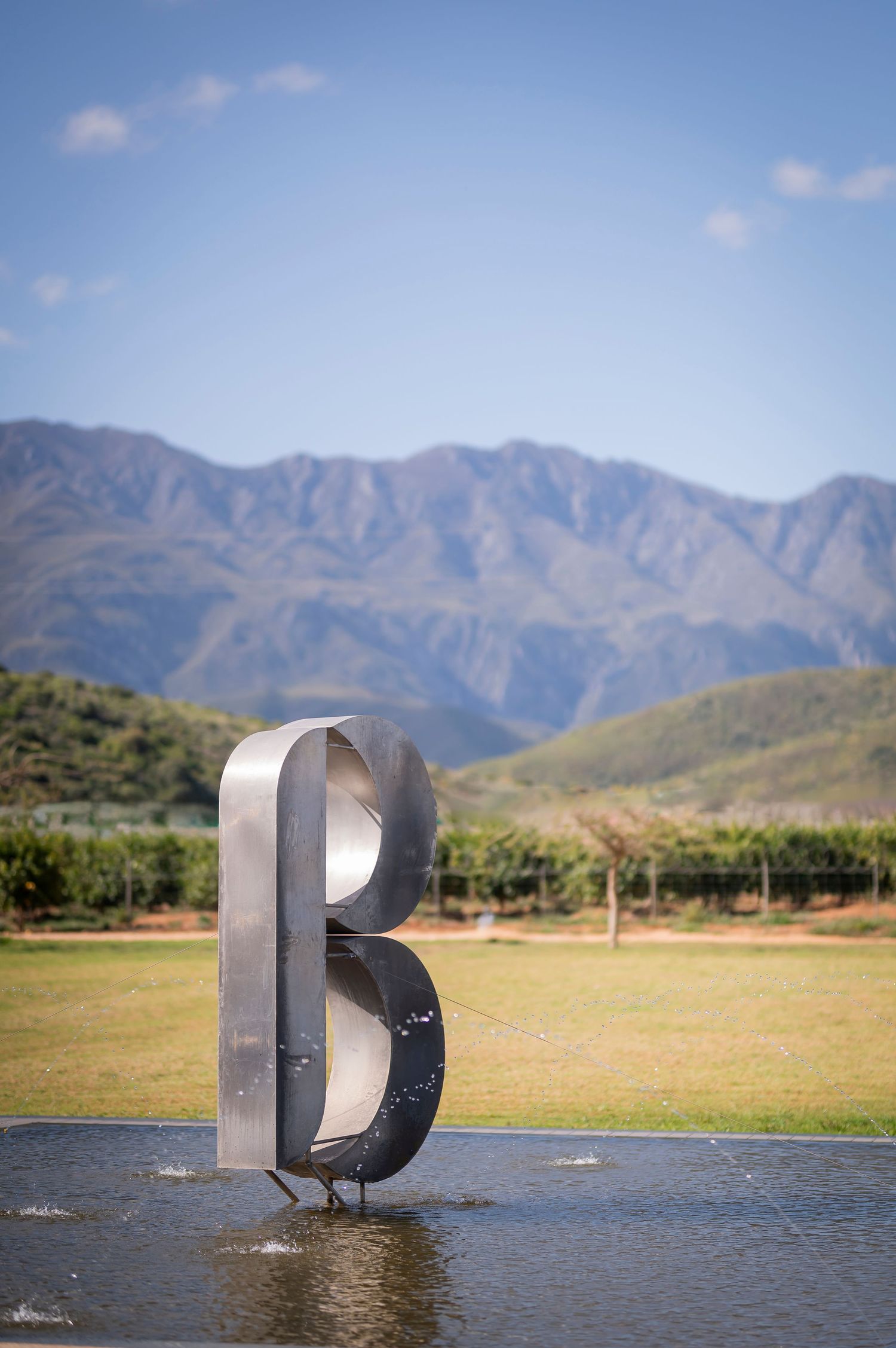 Large metal letter sculpture in shallow water with mountains and blue sky in the background.