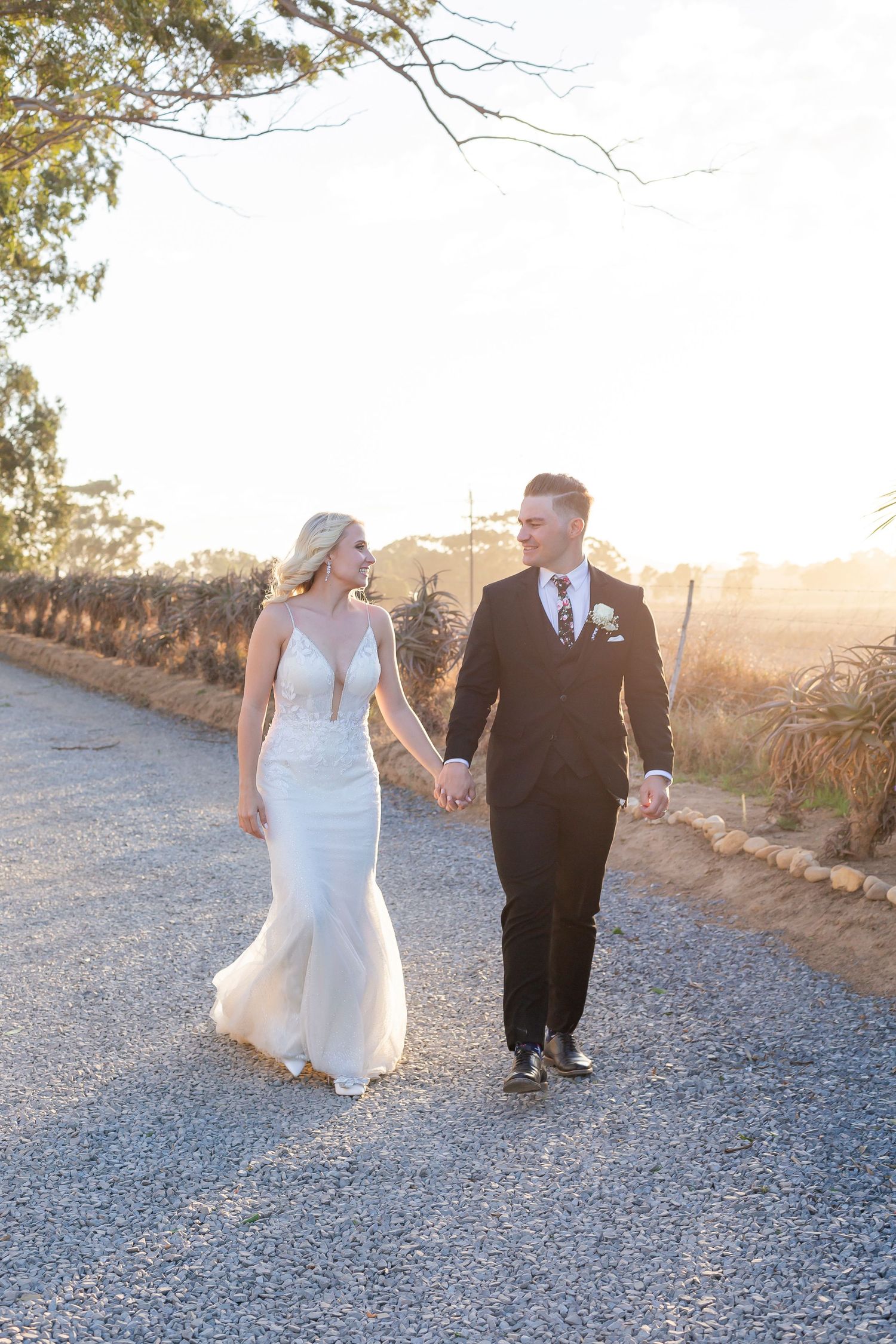 Newlywed couple walks hand in hand along a gravel path at sunset during their romantic outdoor wedding photoshoot.