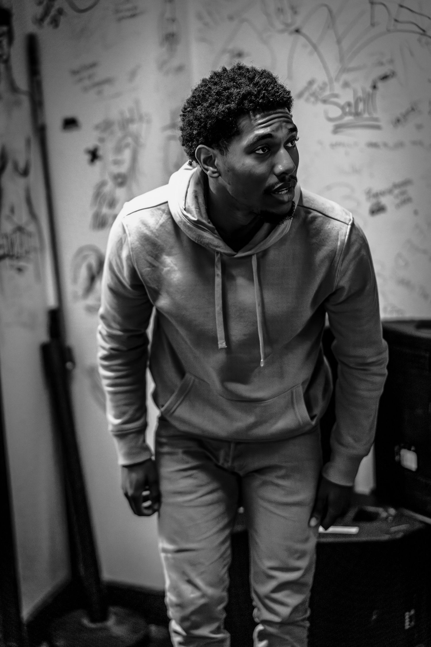 Black and white portrait of a person in a casual grey hoodie and light pants against a wall with graffiti.