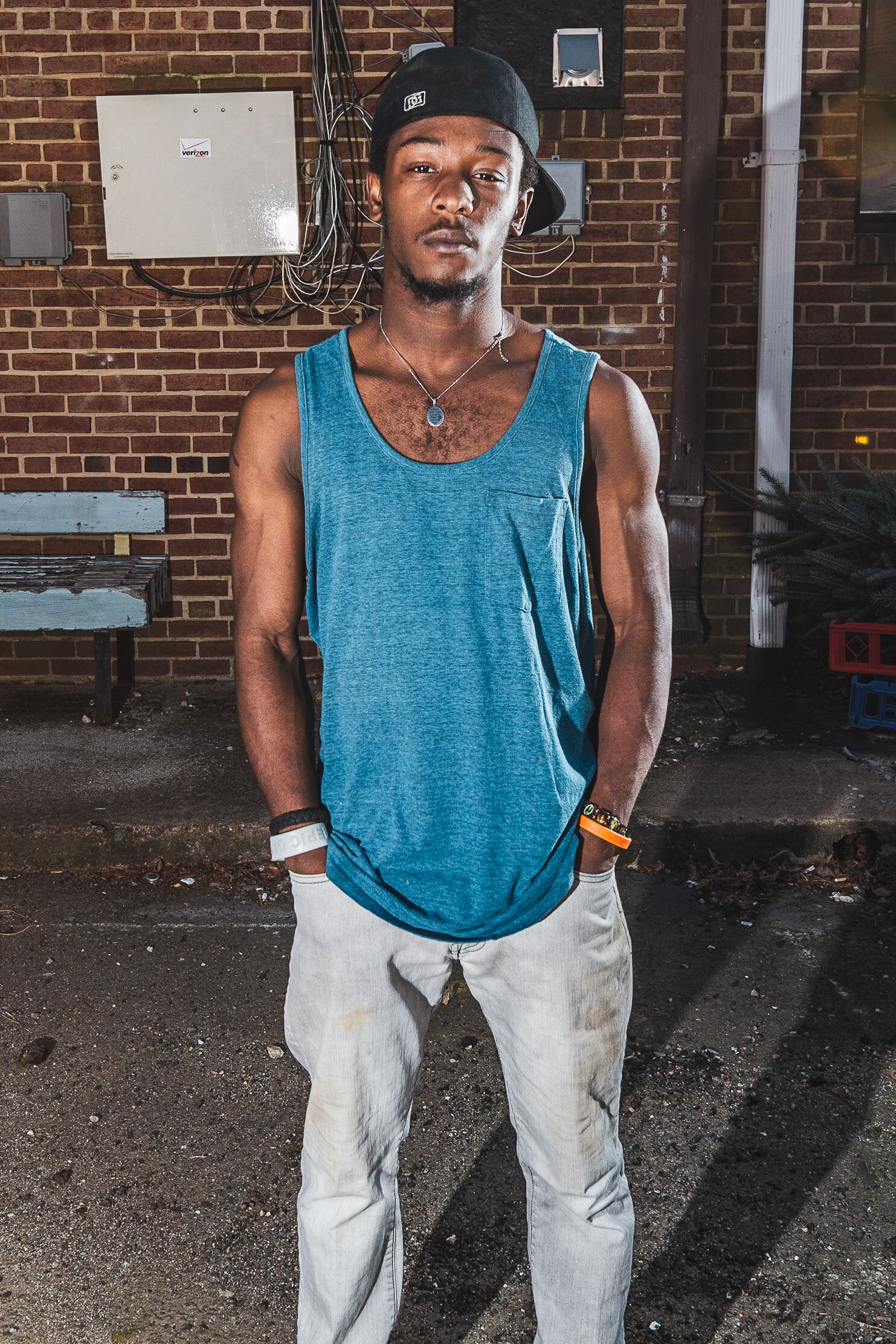 Person standing against brick wall wearing blue tank top, black cap and white pants in urban setting.
