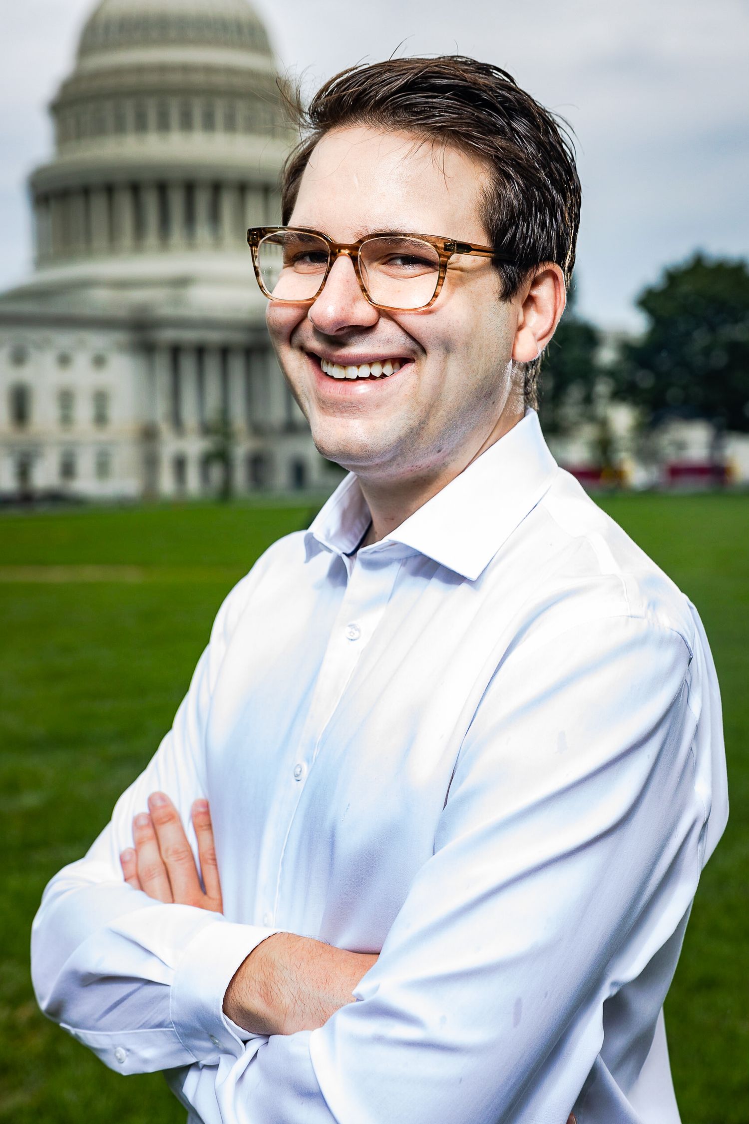 Photo of a person in glasses and white dress shirt smiling with arms crossed in front of the US Capitol building.