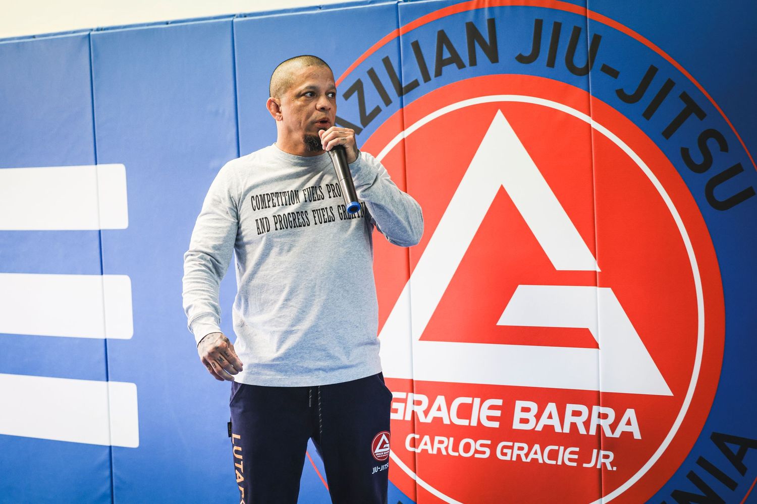 Martial arts instructor in white sweatshirt stands in front of large red Gracie Barra Brazilian Jiu-Jitsu logo.