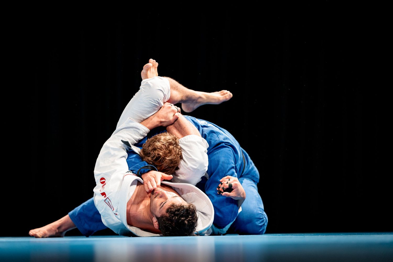 Two martial artists in white and blue gis grappling on a dark mat during competition.