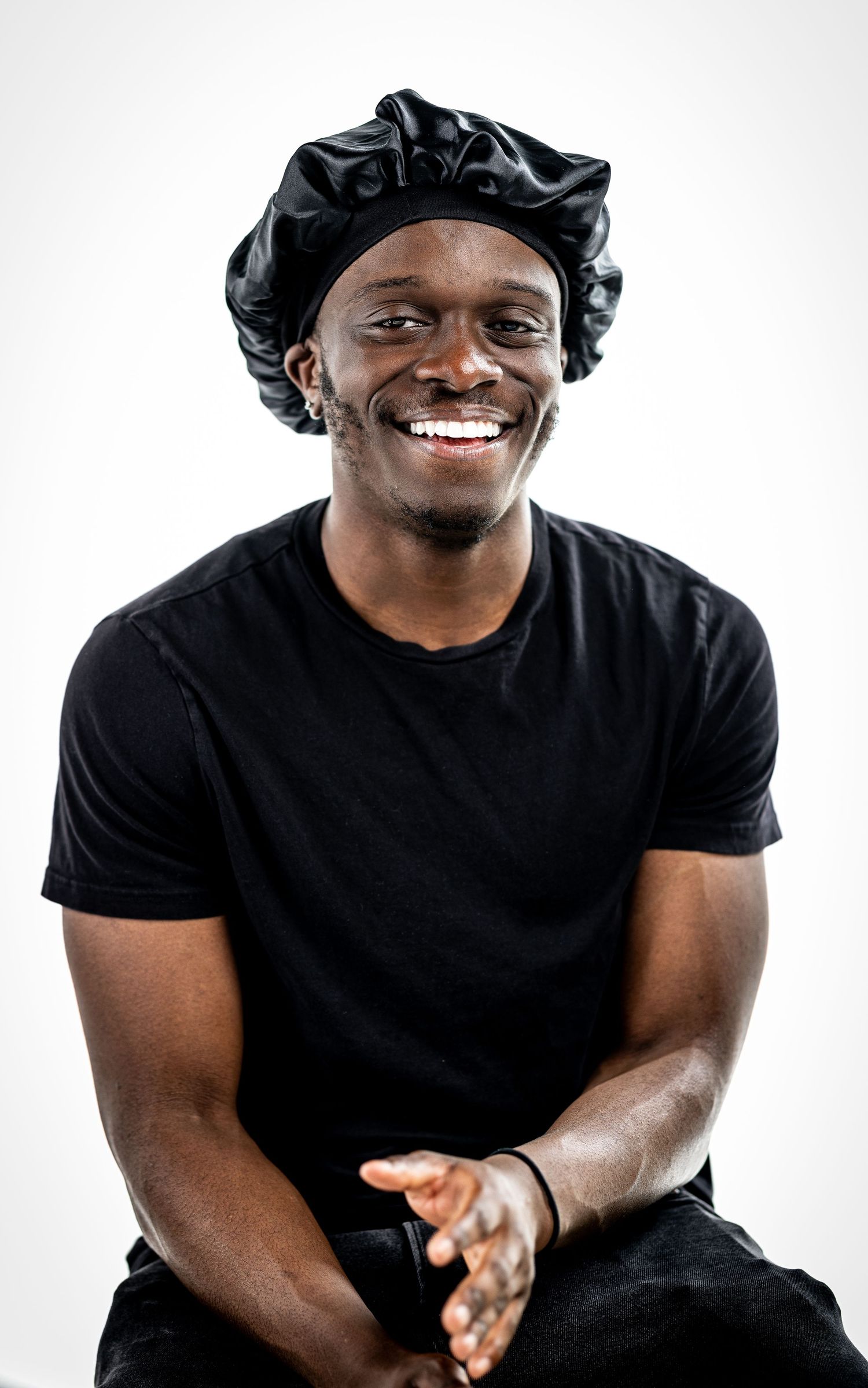 Person wearing black durag and t-shirt smiling warmly against white studio background.