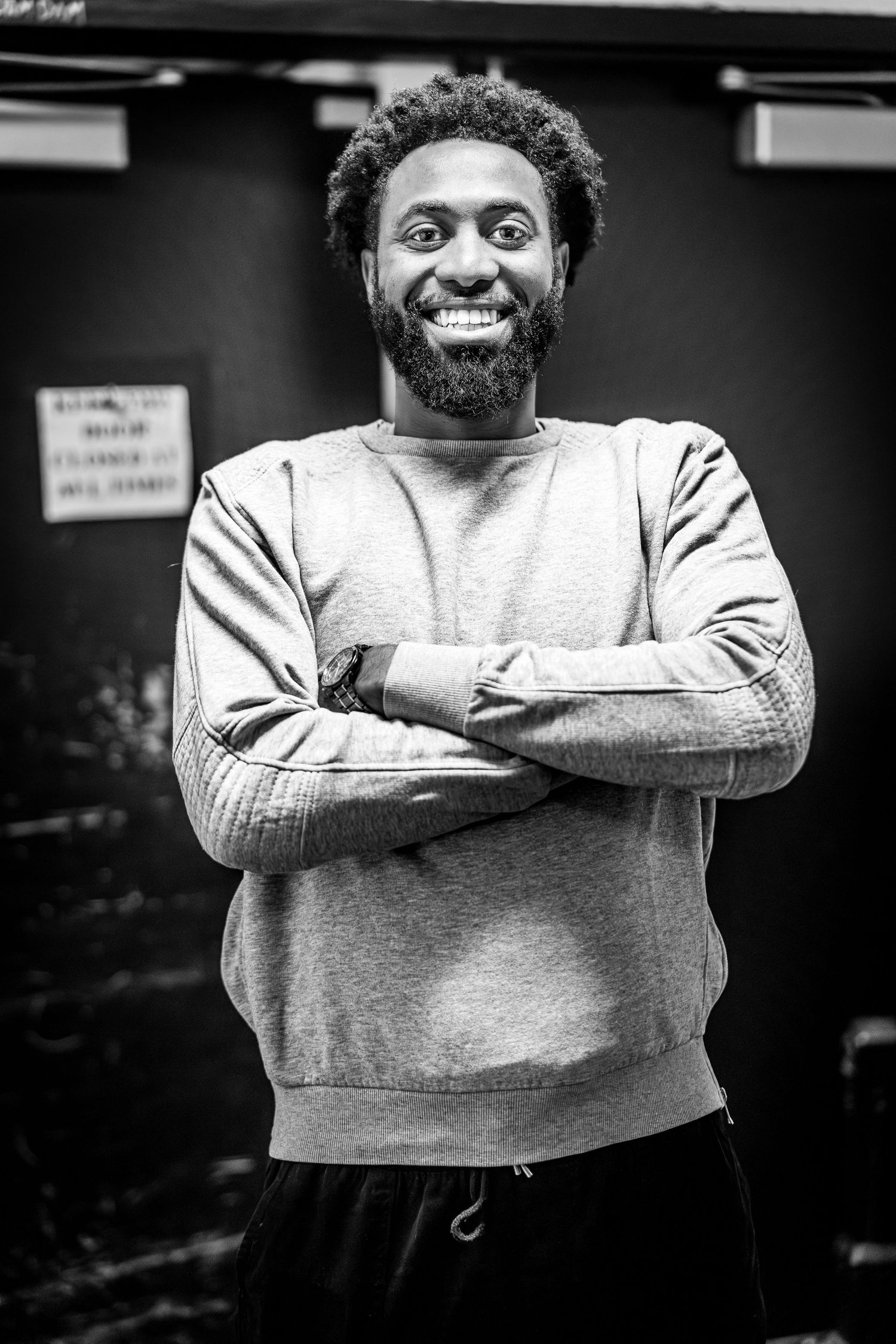 A black and white portrait of a smiling person in a gray sweatshirt with crossed arms against a dark background.