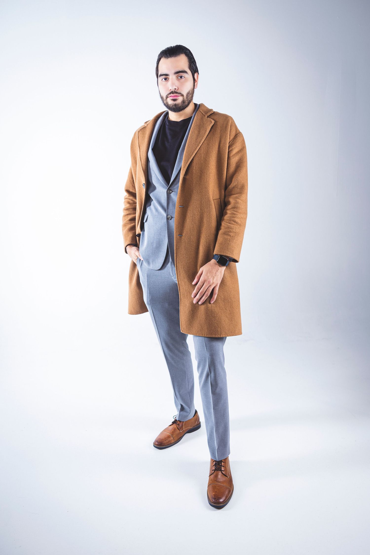 Stylish man in camel coat and gray suit poses against white background.