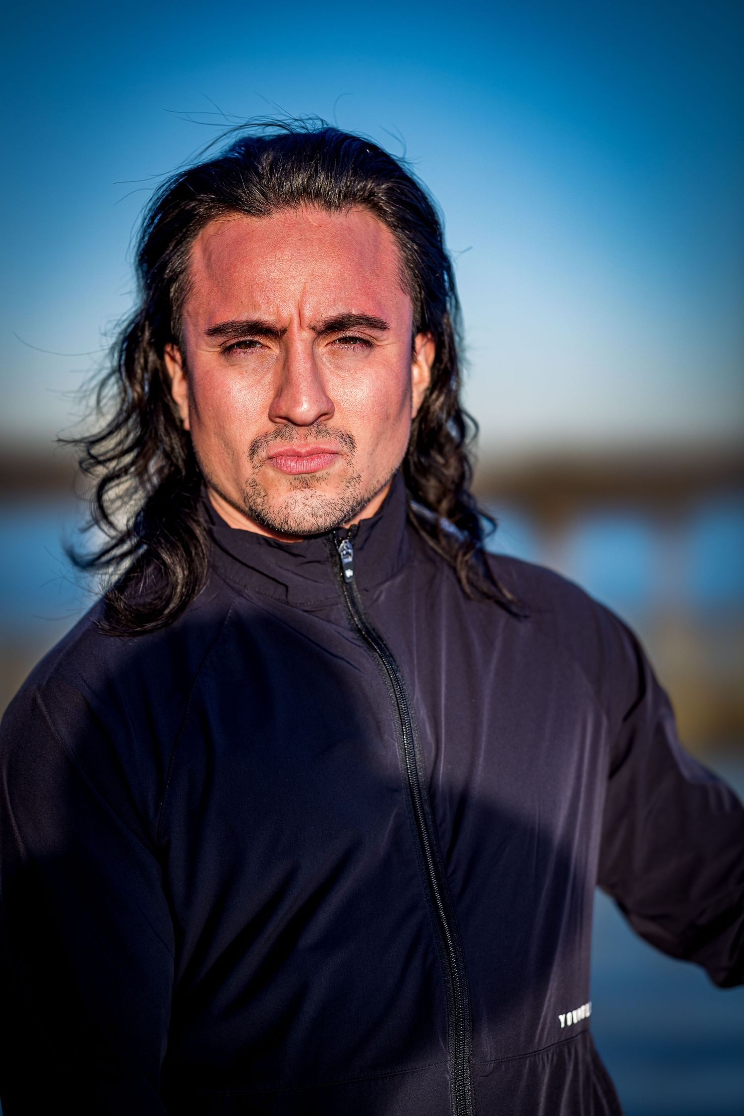 Athletic figure in black zip-up jacket photographed outdoors against blue sky during golden hour lighting.
