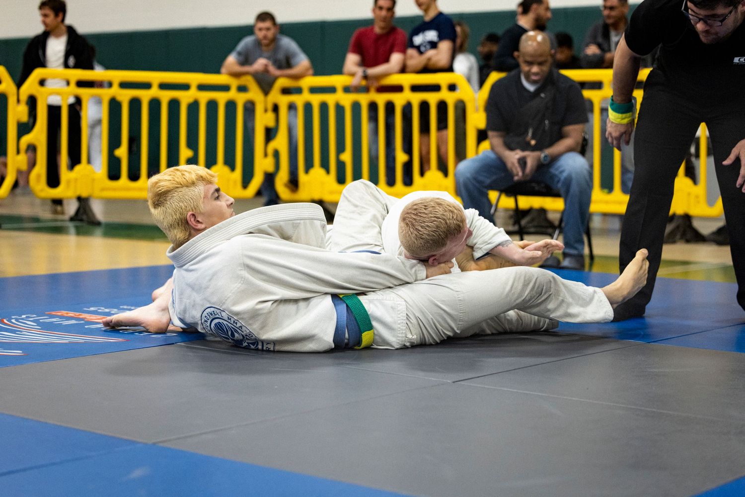 Two martial artists in white gis grapple during a Brazilian jiu-jitsu competition match on blue mats.