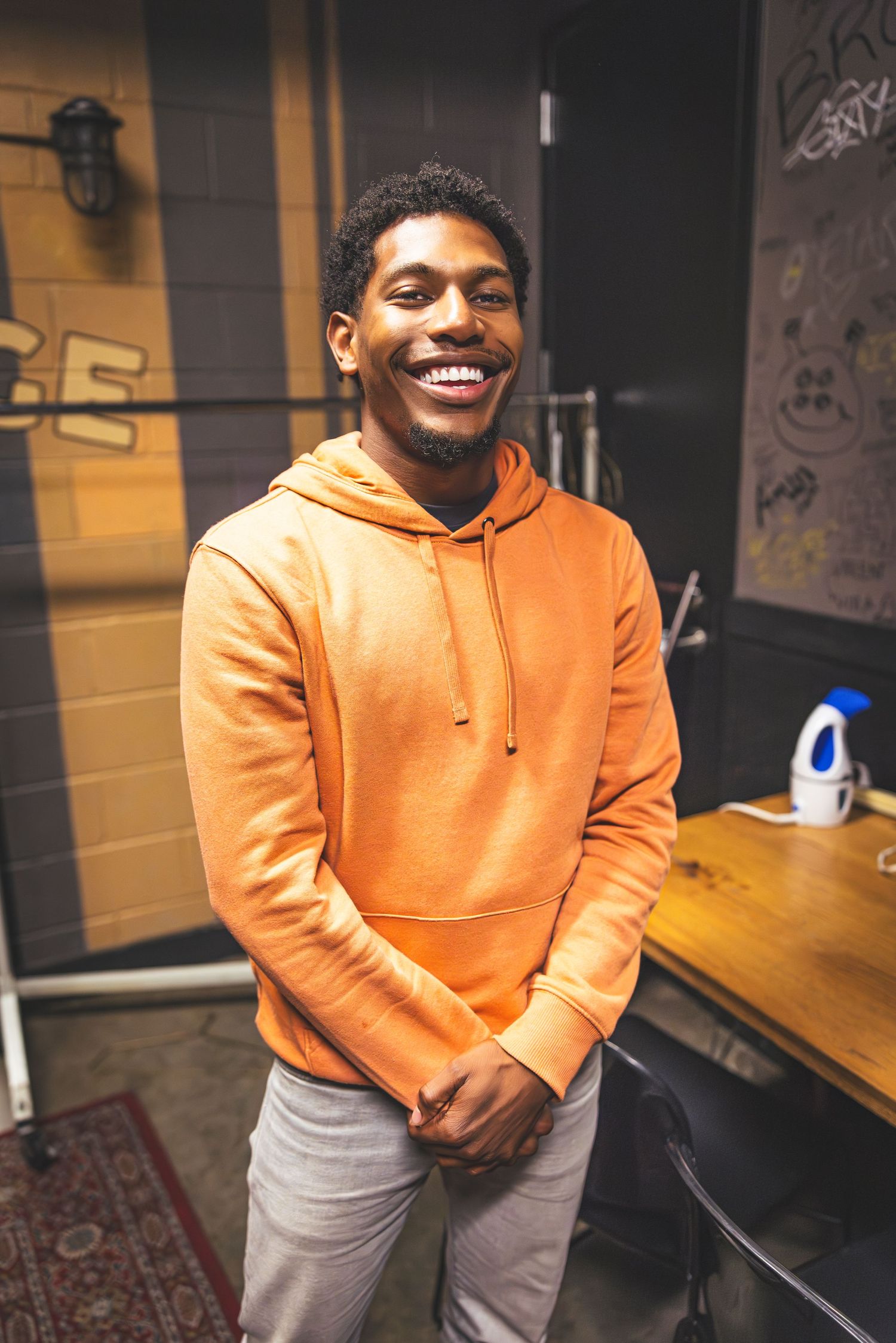 A person wearing an orange hoodie and light gray pants smiles warmly while standing in a casual indoor setting.