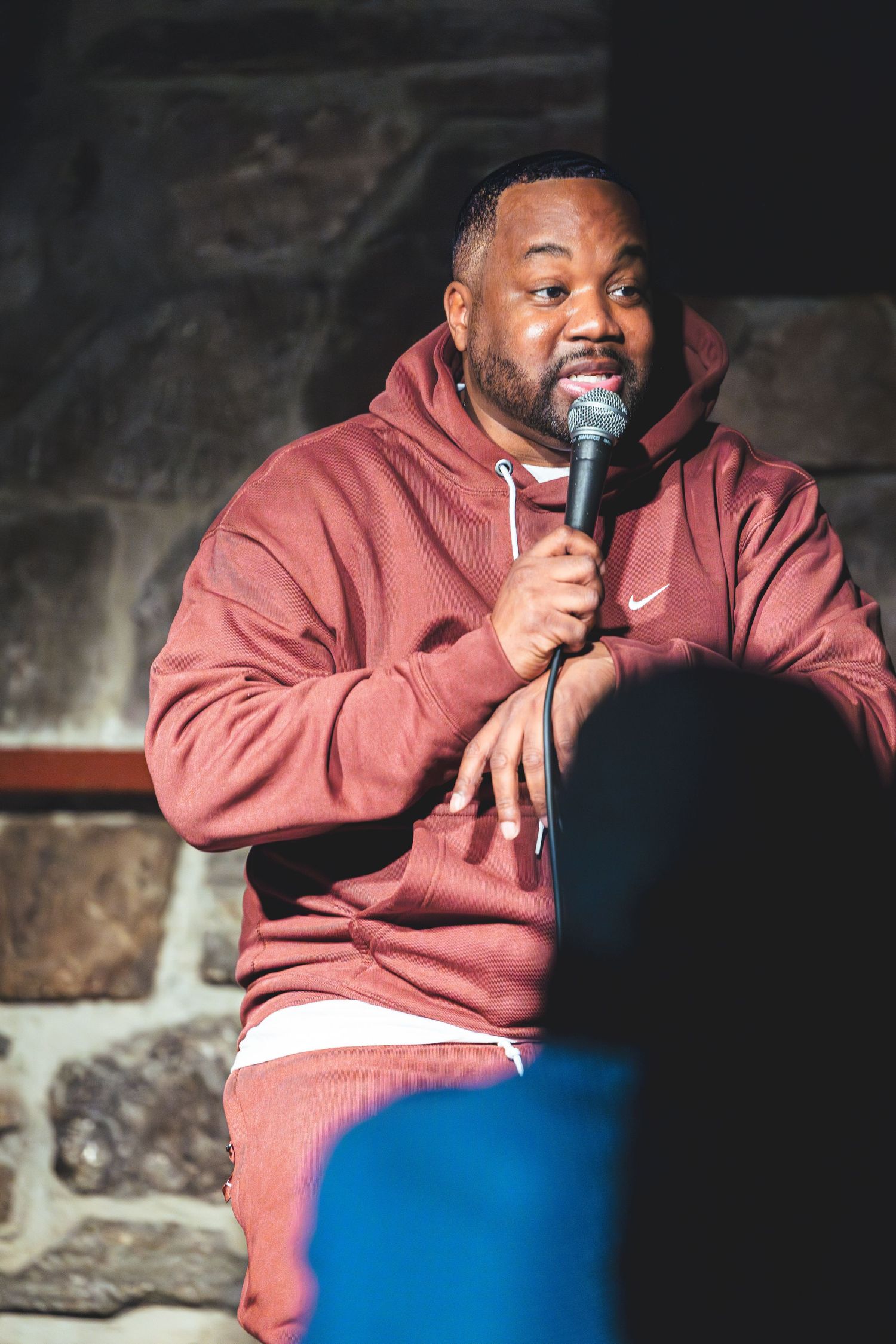 Performer in a burgundy Nike hoodie holds microphone while seated on stage during comedy show.