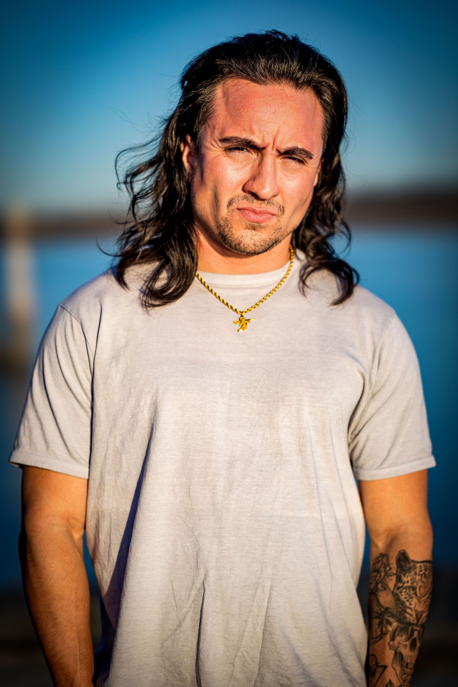 Portrait of person with long dark hair wearing white t-shirt and gold necklace against blurred waterfront background at sunset.