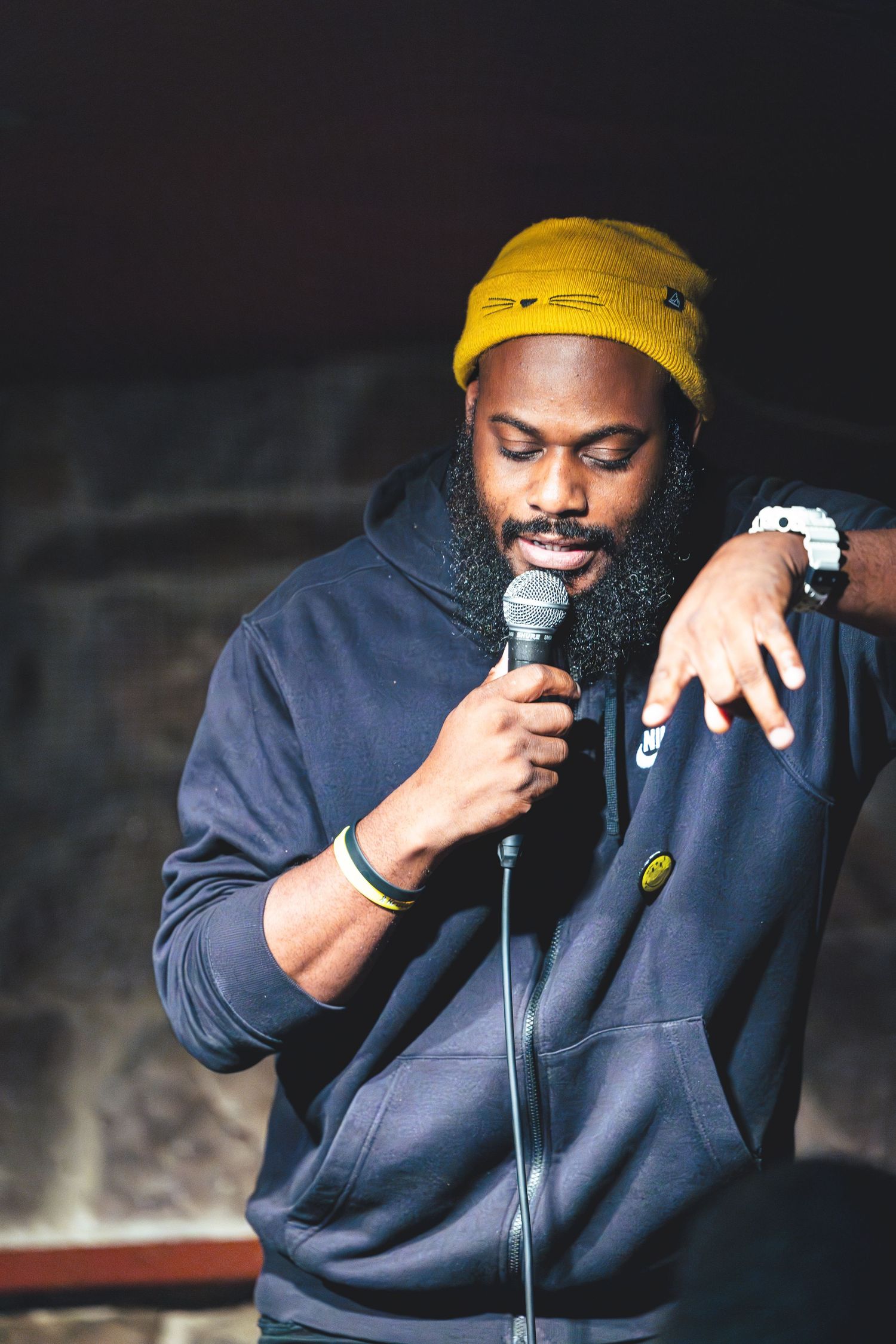 A performer wearing a yellow beanie and navy hoodie speaks into a microphone during a stand-up comedy performance.