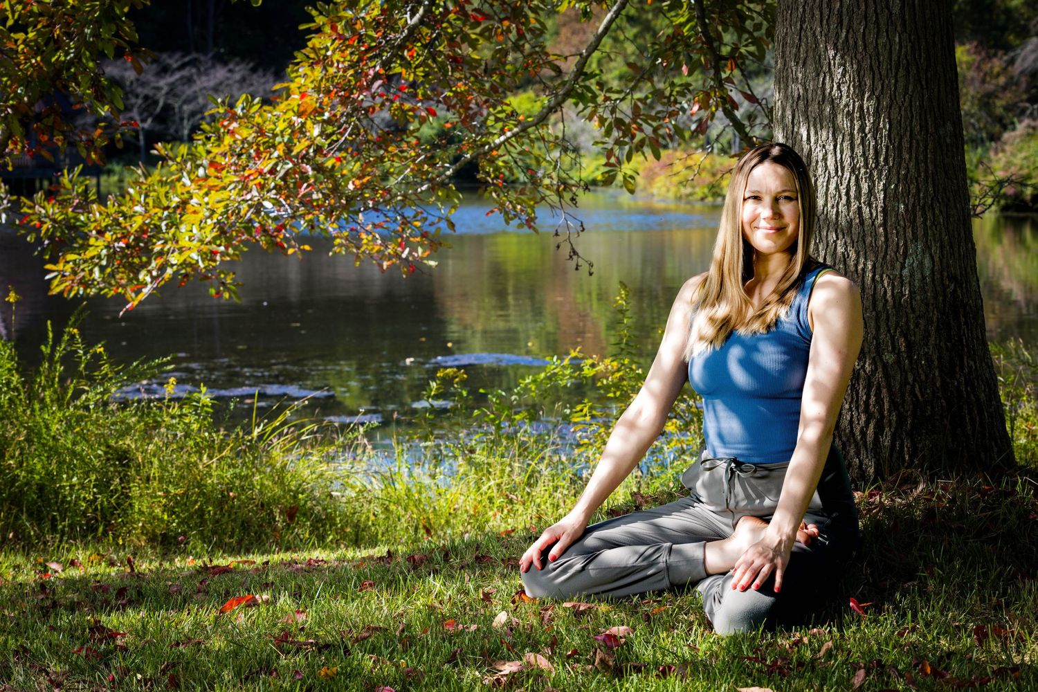 A person in a blue dress rests against a large tree trunk by a lake with autumn foliage in the background.