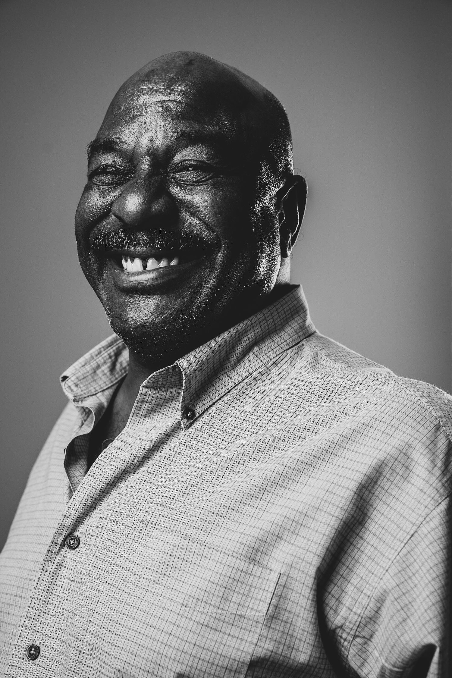 Black and white portrait photograph of a joyful person wearing a plaid button-up shirt against a gradient background.