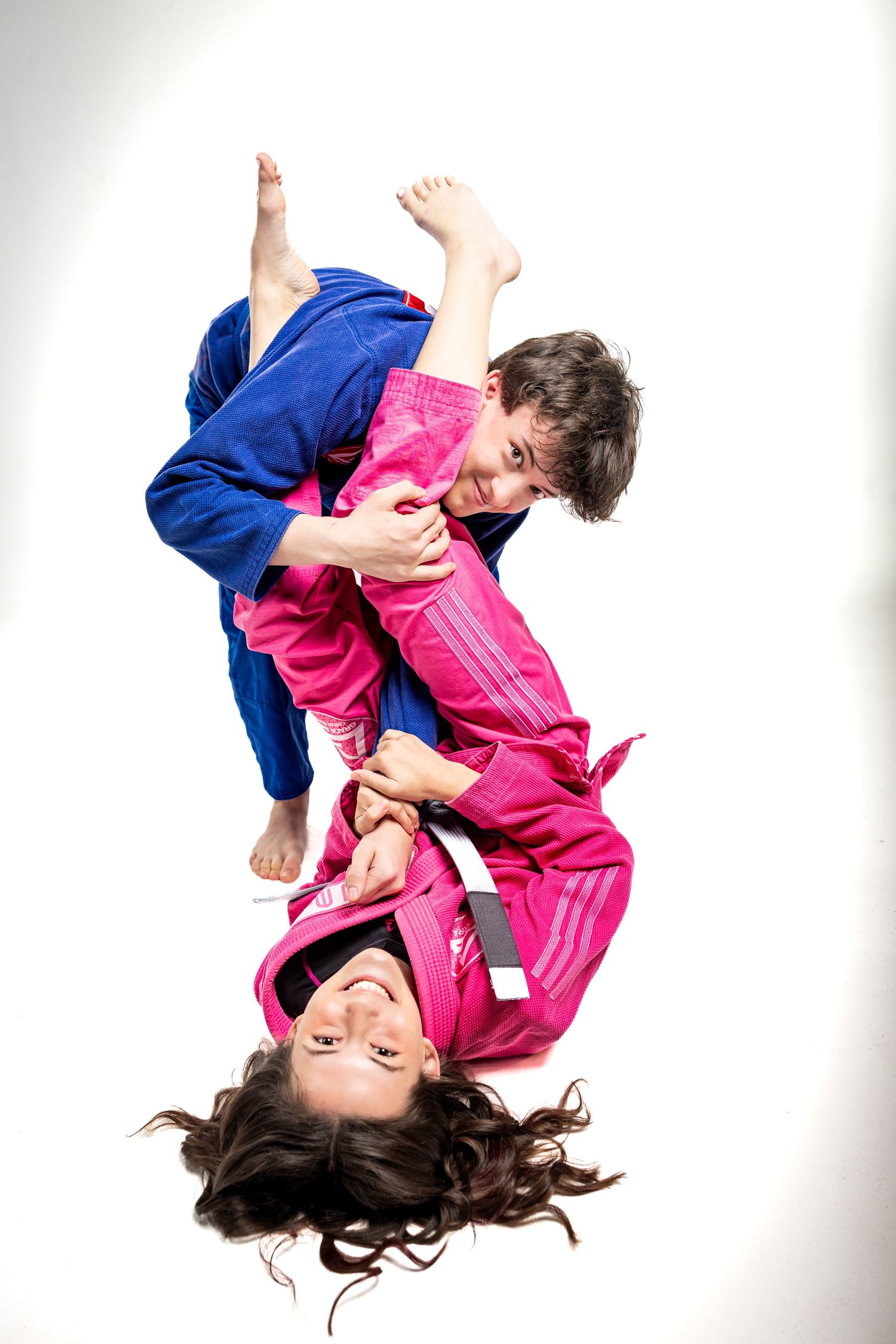 Two martial artists in pink and blue gis practicing grappling moves on a white background.