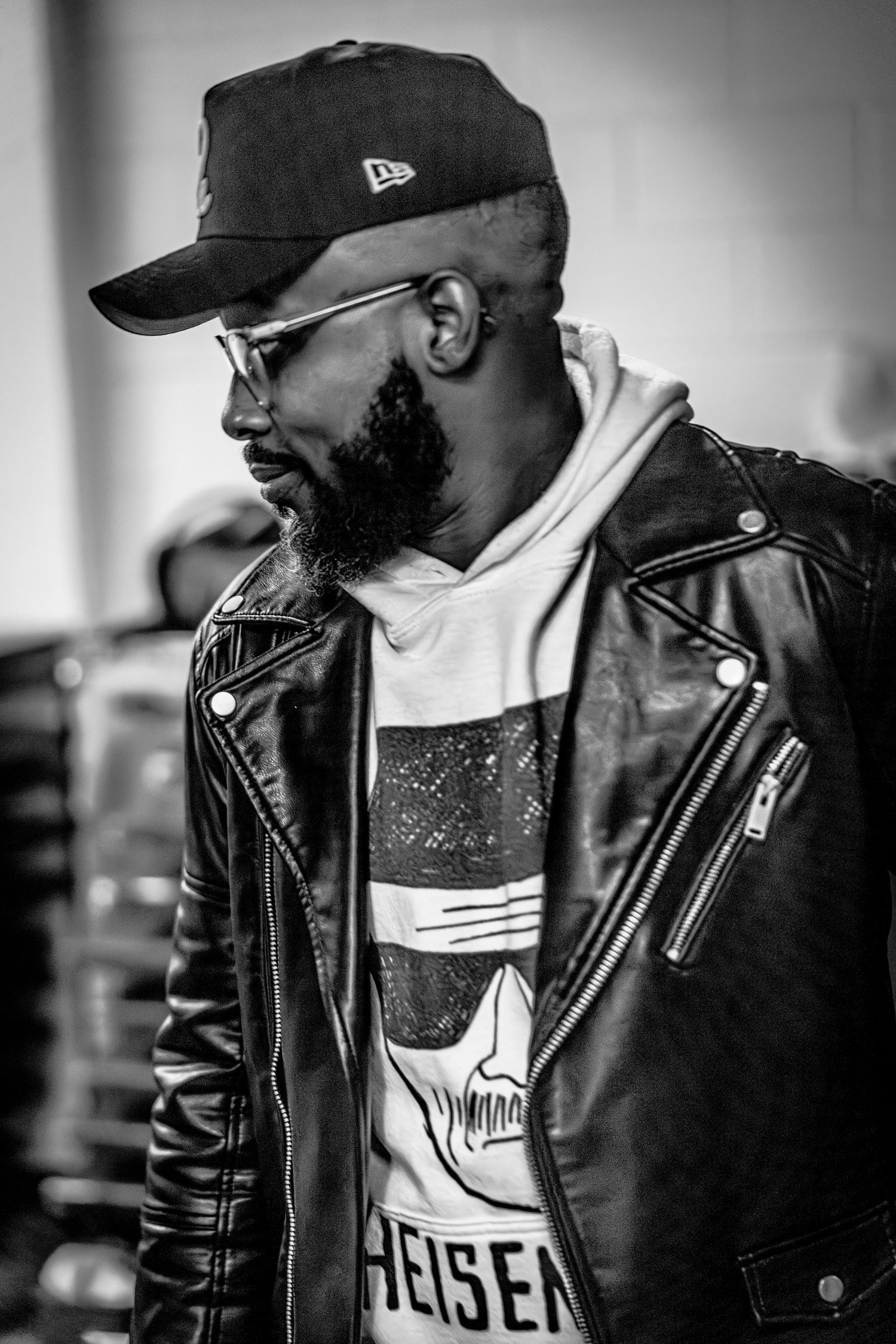 Black and white photo showing person in leather jacket, New Era cap and glasses in profile view against blurred background.