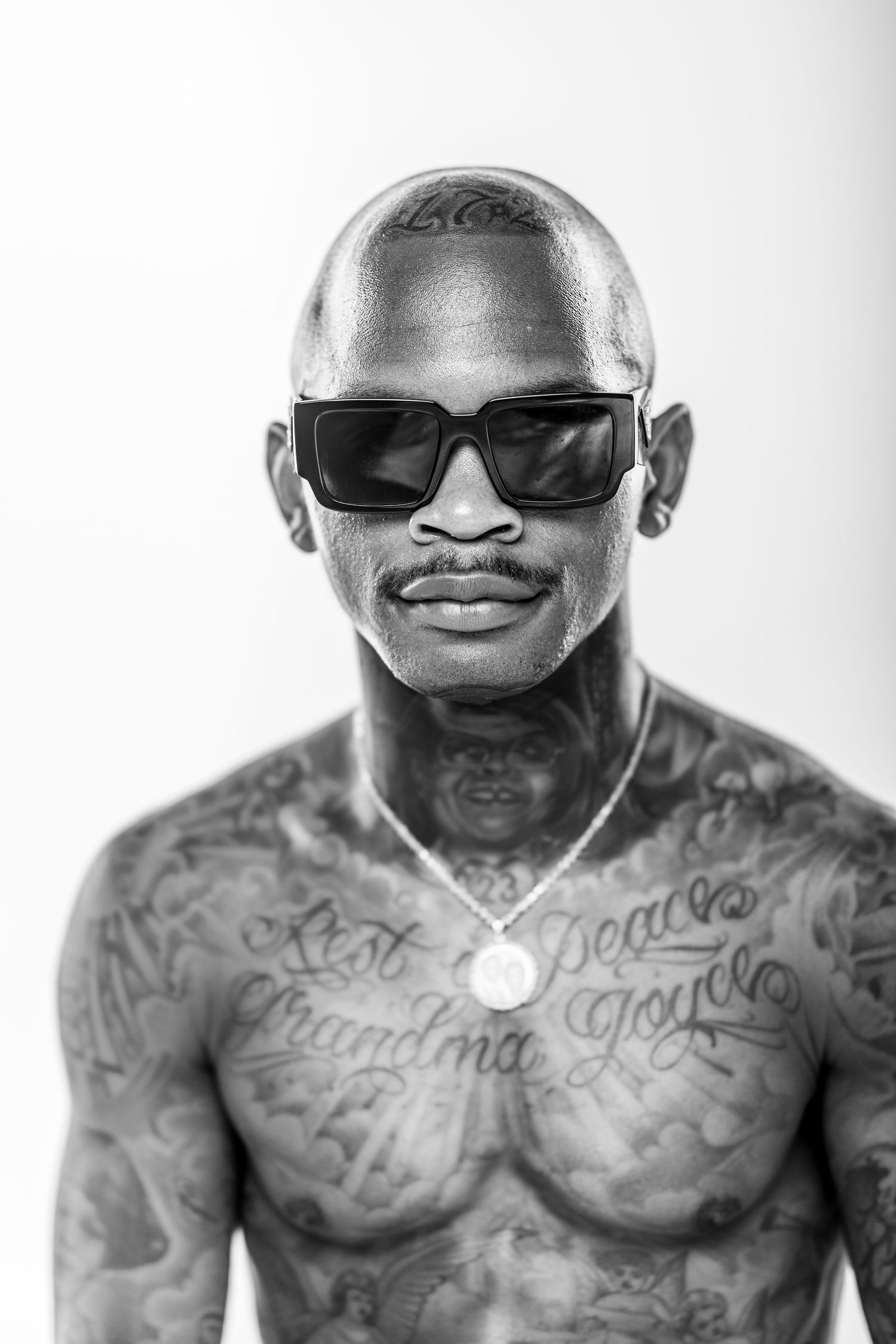 Black and white portrait of a muscular man with sunglasses and chest tattoos posing shirtless against white background.