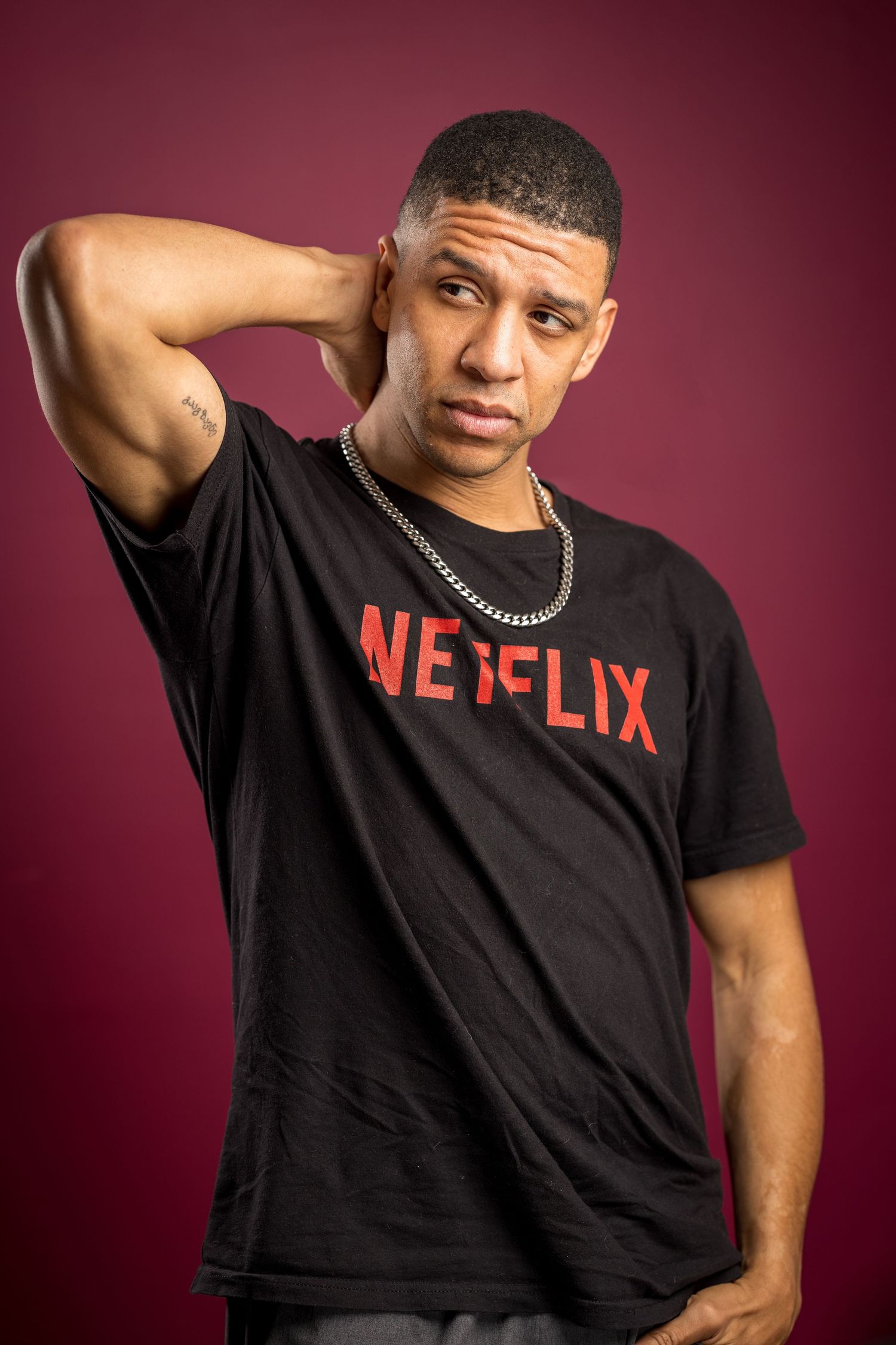 Someone wearing a black Netflix t-shirt poses with hand behind head against burgundy background.