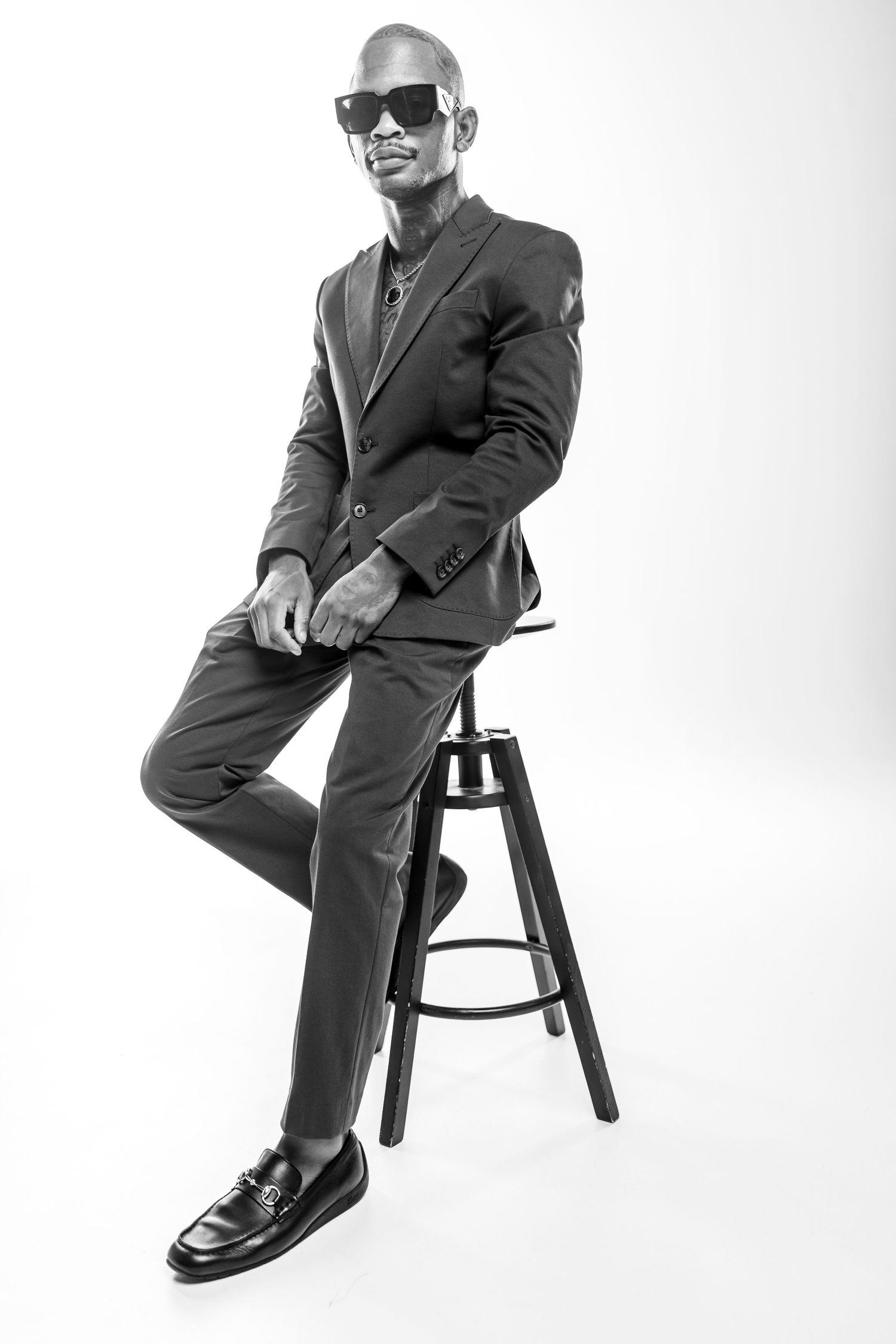 Stylish portrait in black and white showing someone in a suit and sunglasses sitting confidently on a bar stool.