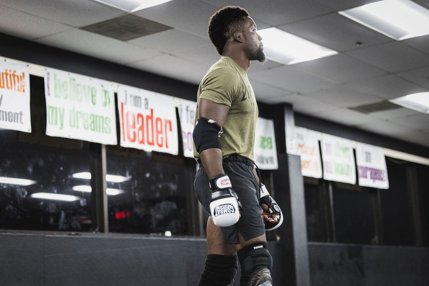 A fighter wearing knee pads and a yellow t-shirt stands in a gym with promotional banners hanging on the wall.