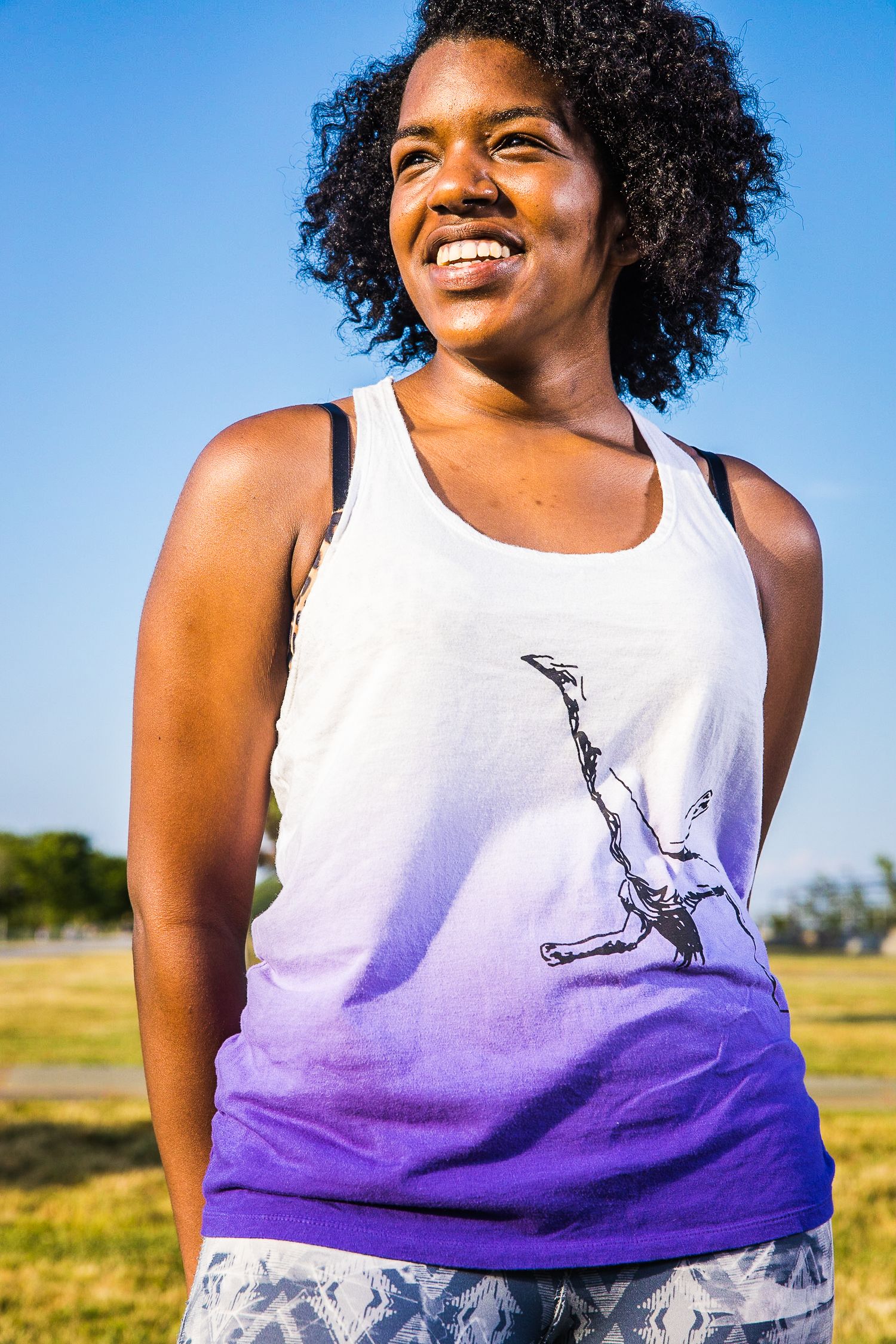 A person wearing an ombre white to purple tank top with a dancer silhouette design smiles outdoors on a sunny day.
