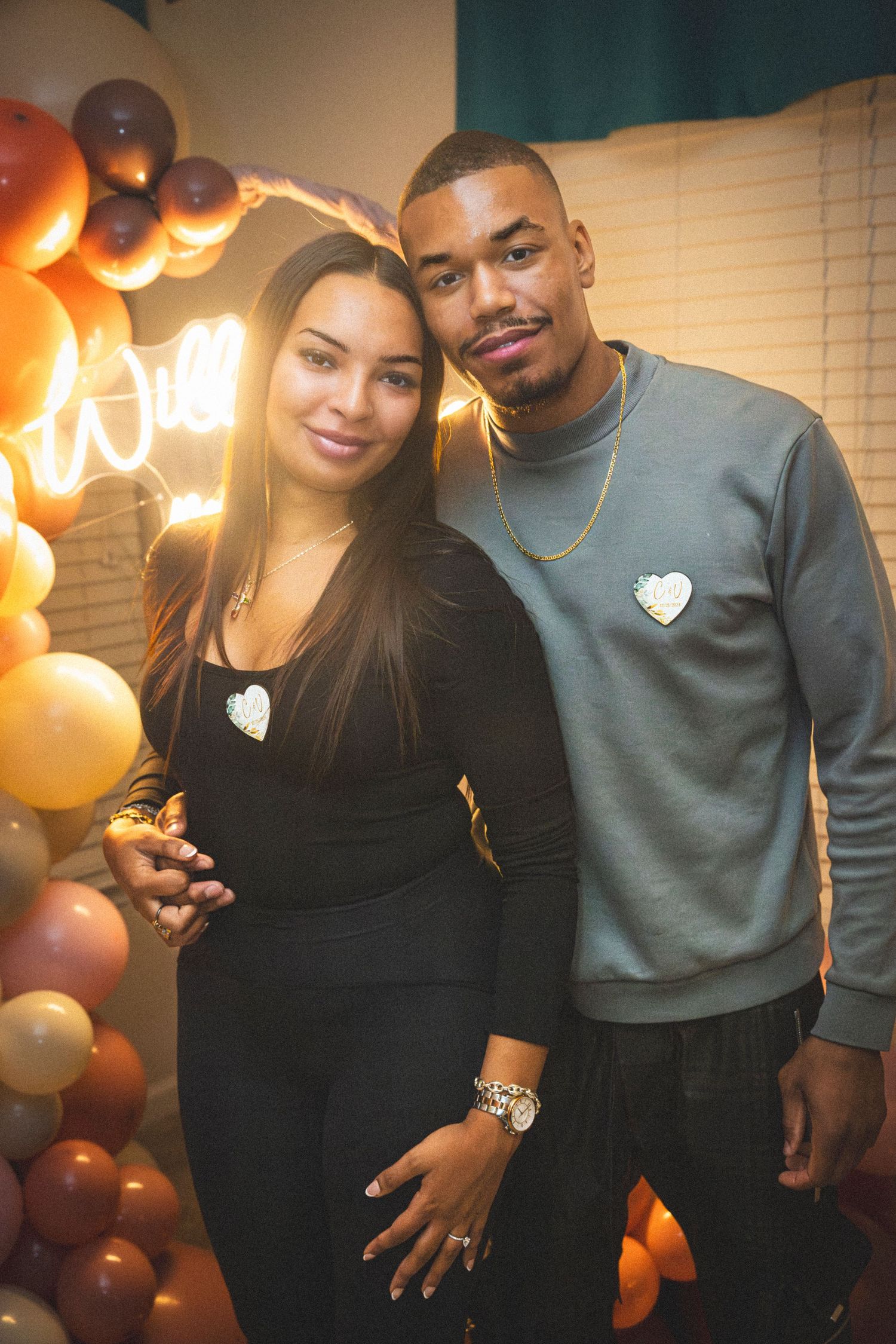 A couple wearing matching heart sweaters poses in front of copper balloon arch and neon lights.