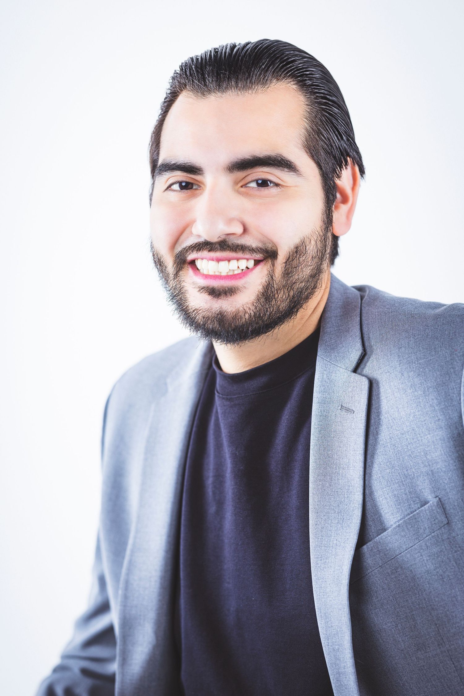 Professional headshot of person in grey blazer and black shirt smiling warmly.